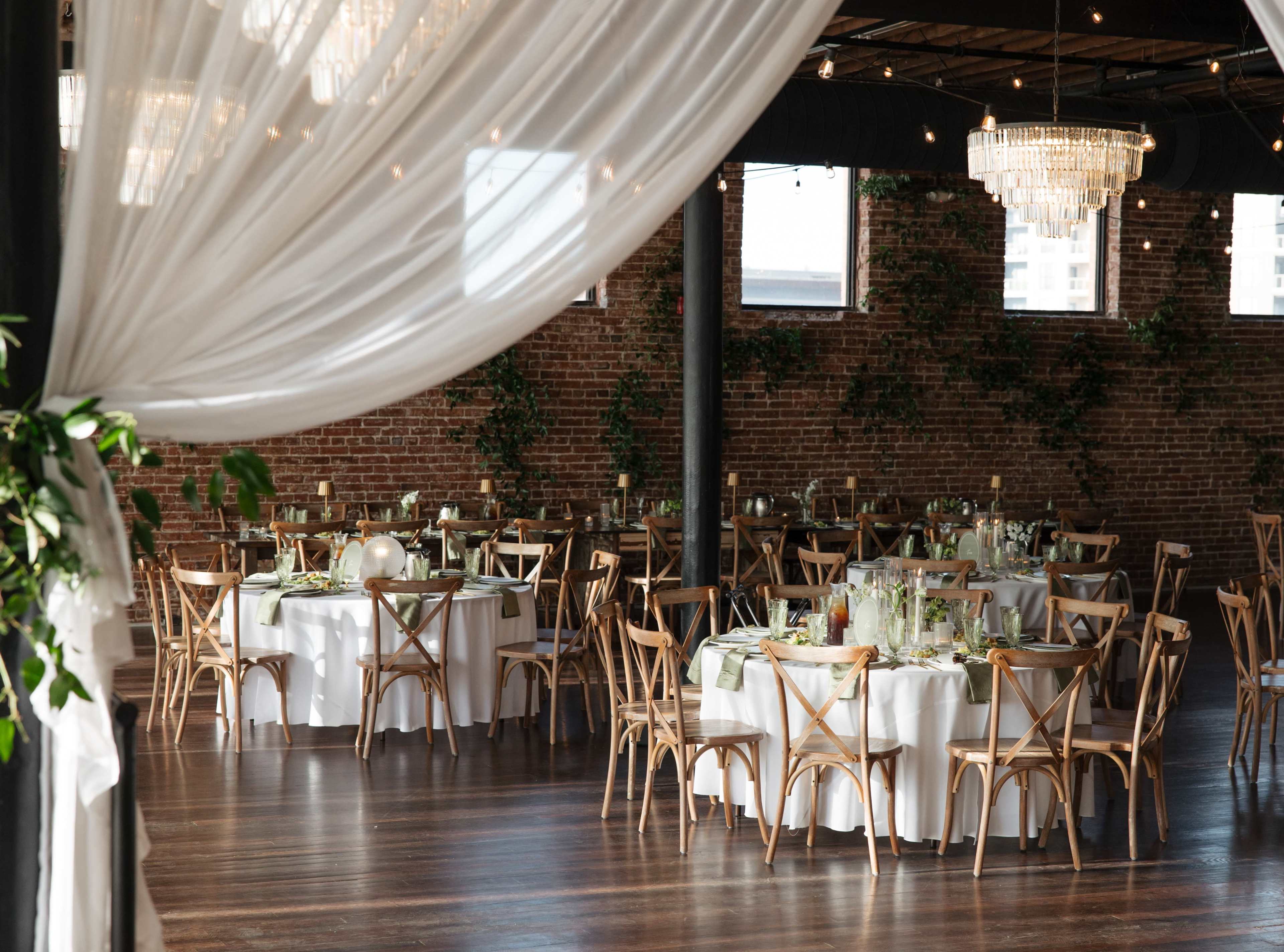 A dining hall with round tables set for a formal event, featuring white tablecloths, wooden chairs, and decorative centerpieces, under soft lighting and hanging chandeliers.