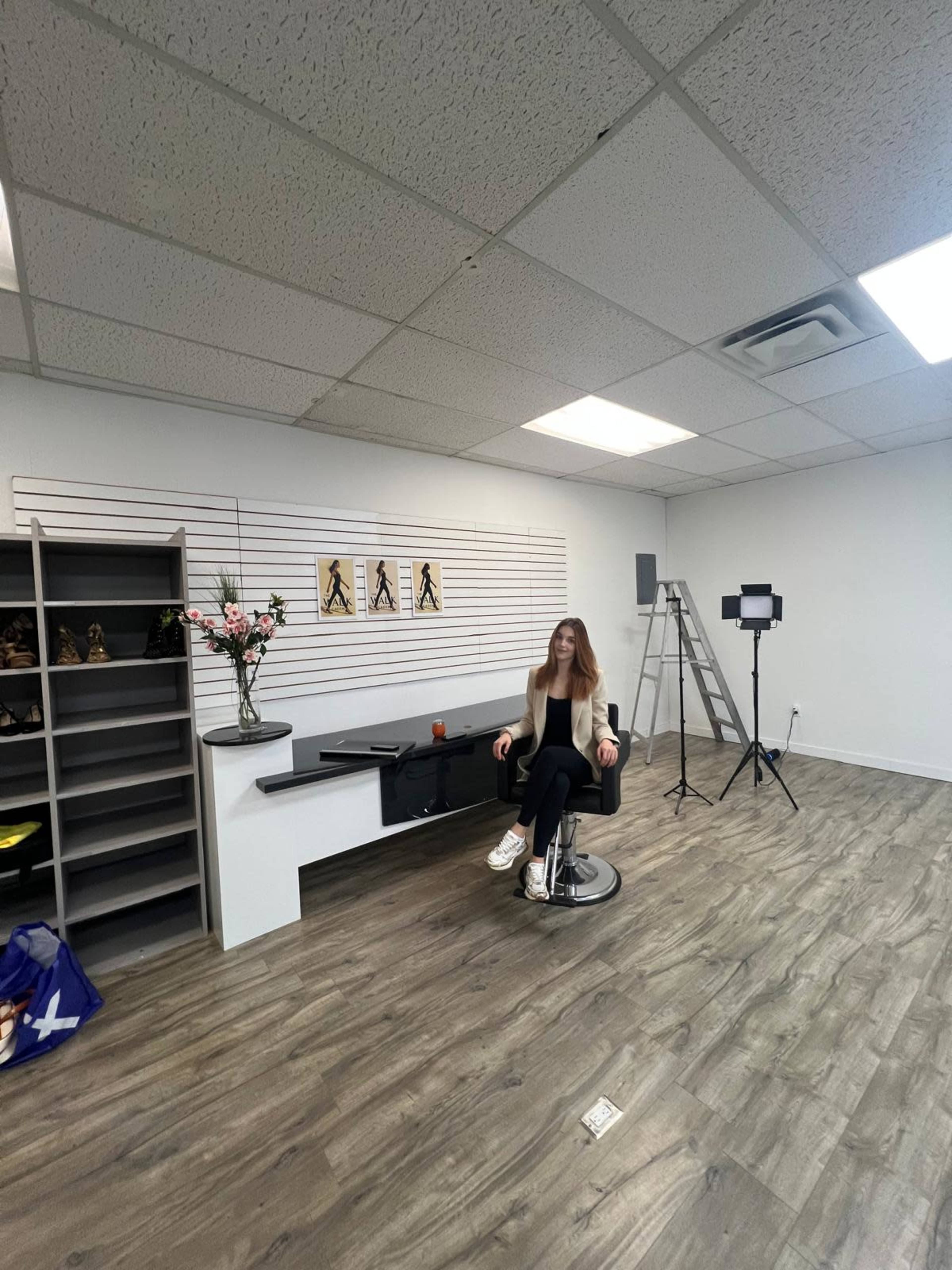 A woman sits in a salon chair in a minimalistic beauty studio with shelves, a ladder, and lighting equipment in the background.