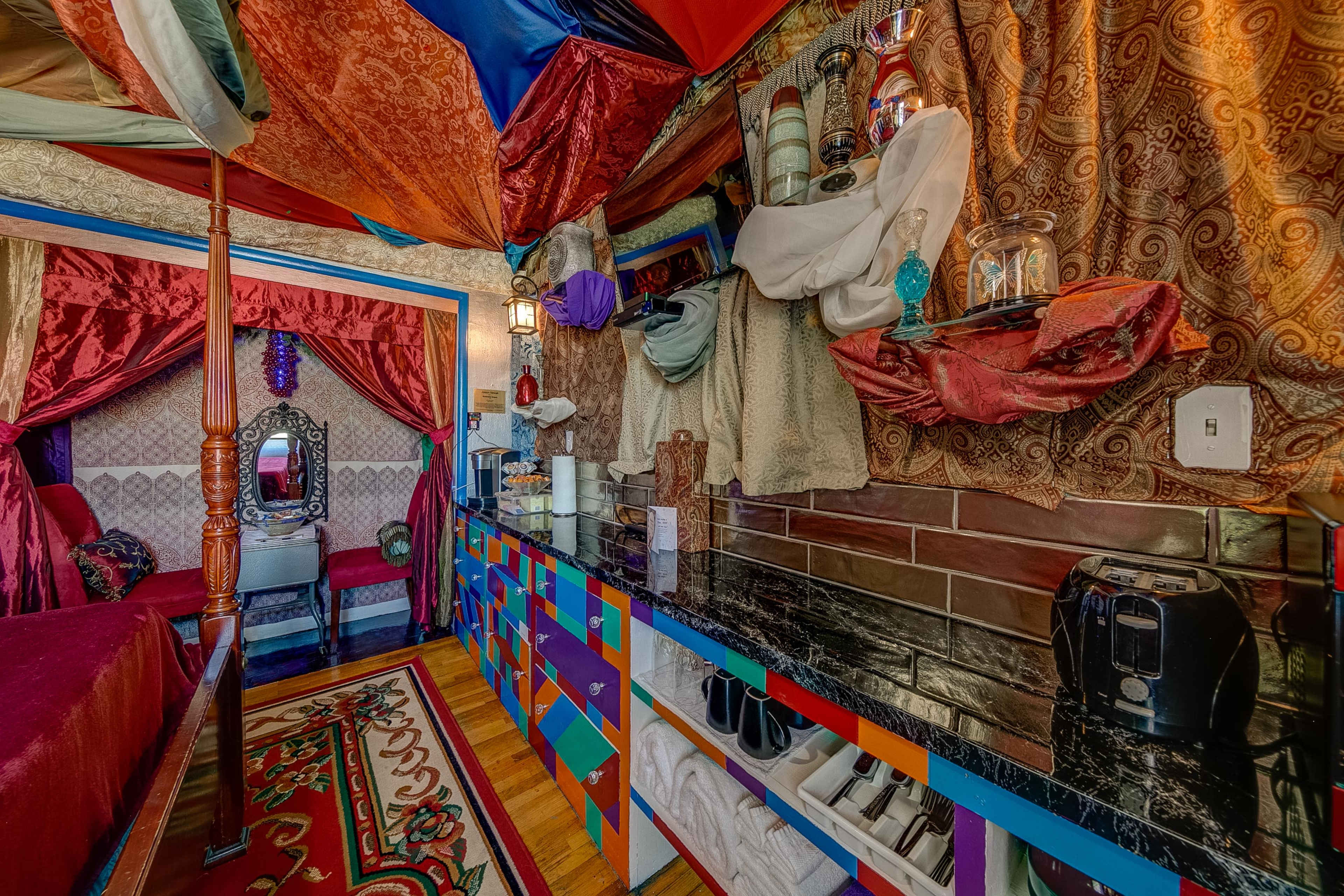 The image shows a vibrant room with a colorful, draped ceiling above a bed and a decorative mirror, flanked by a wall with varied textures and a counter displaying kitchen appliances.