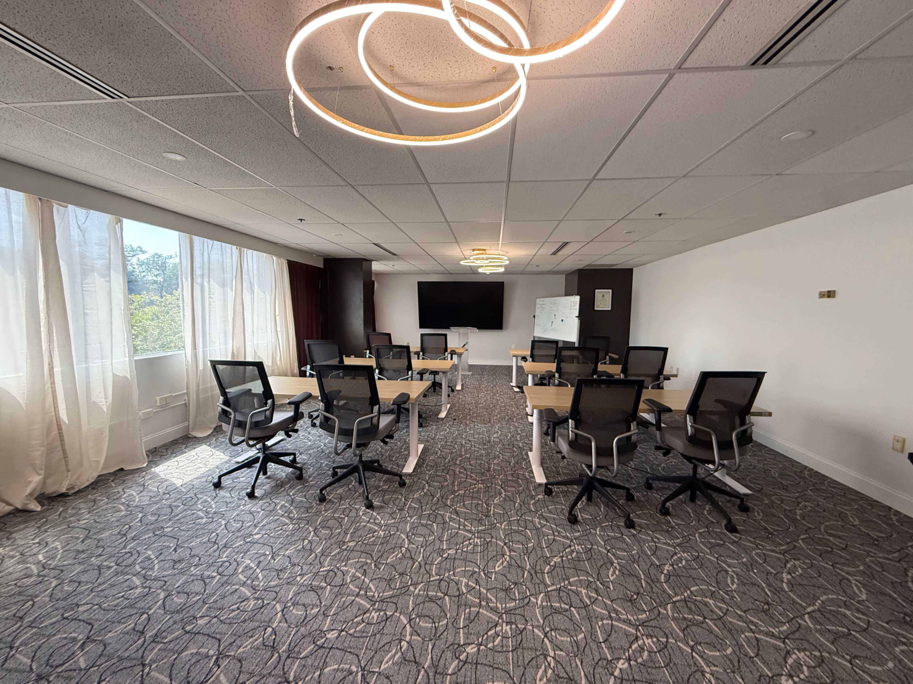 A modern conference room features multiple desks with ergonomic chairs, large windows allowing natural light, and a central light fixture.