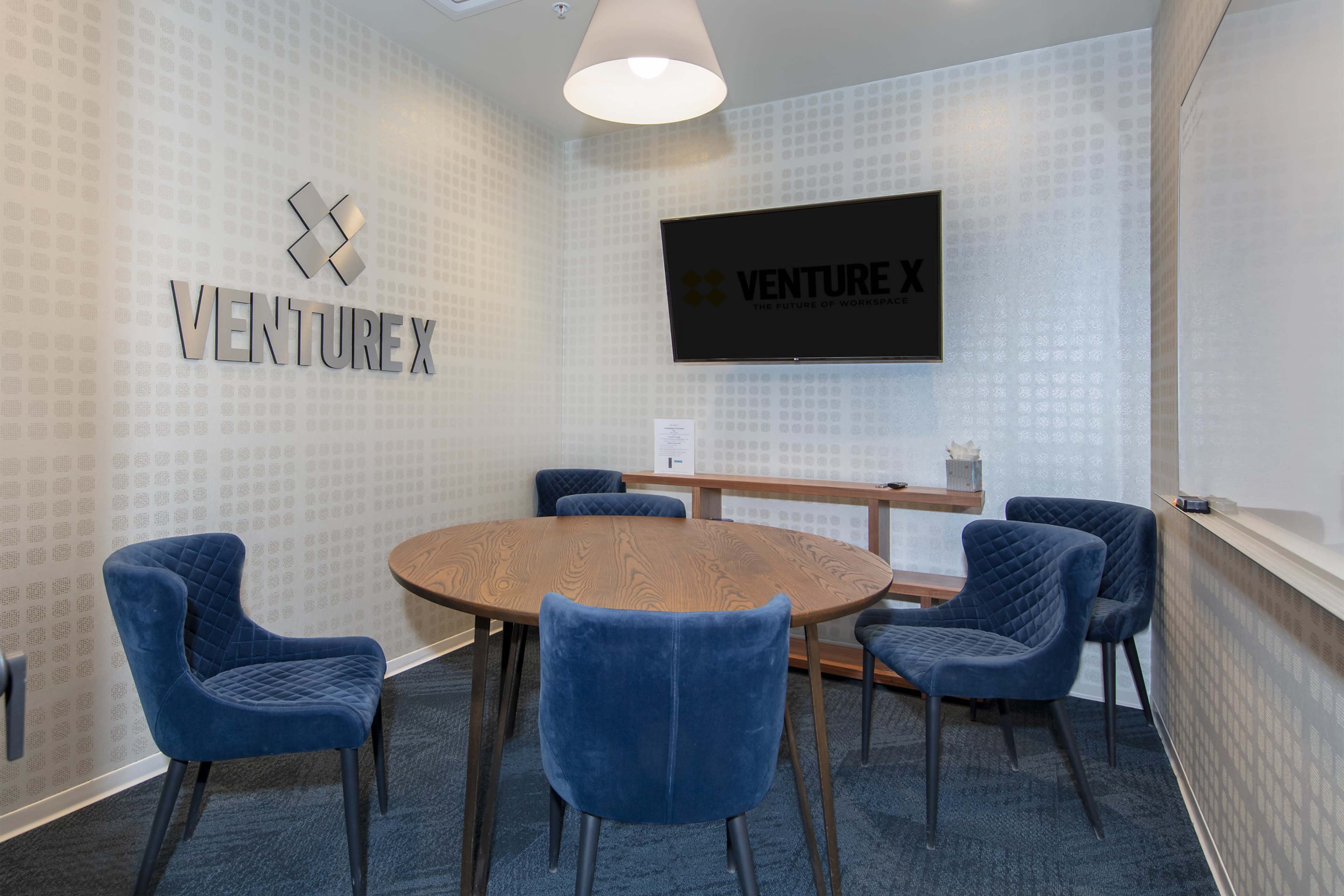 A modern conference room featuring a round wooden table surrounded by four blue upholstered chairs, with a large screen mounted on the wall displaying the "VENTURE X" logo.