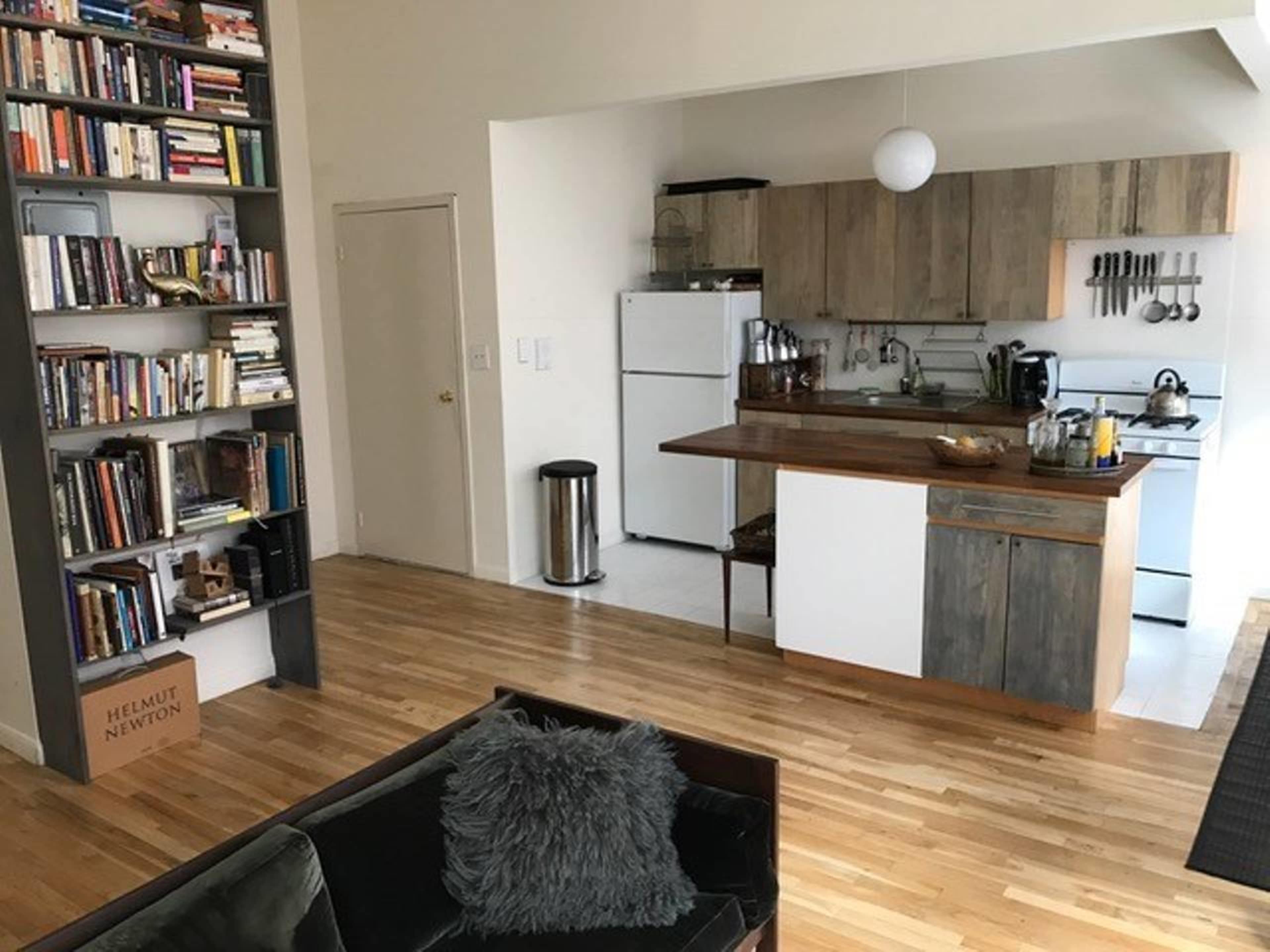 The image shows a living space featuring a kitchen with wooden countertops, a bookshelf filled with books, and a sitting area with a dark couch.