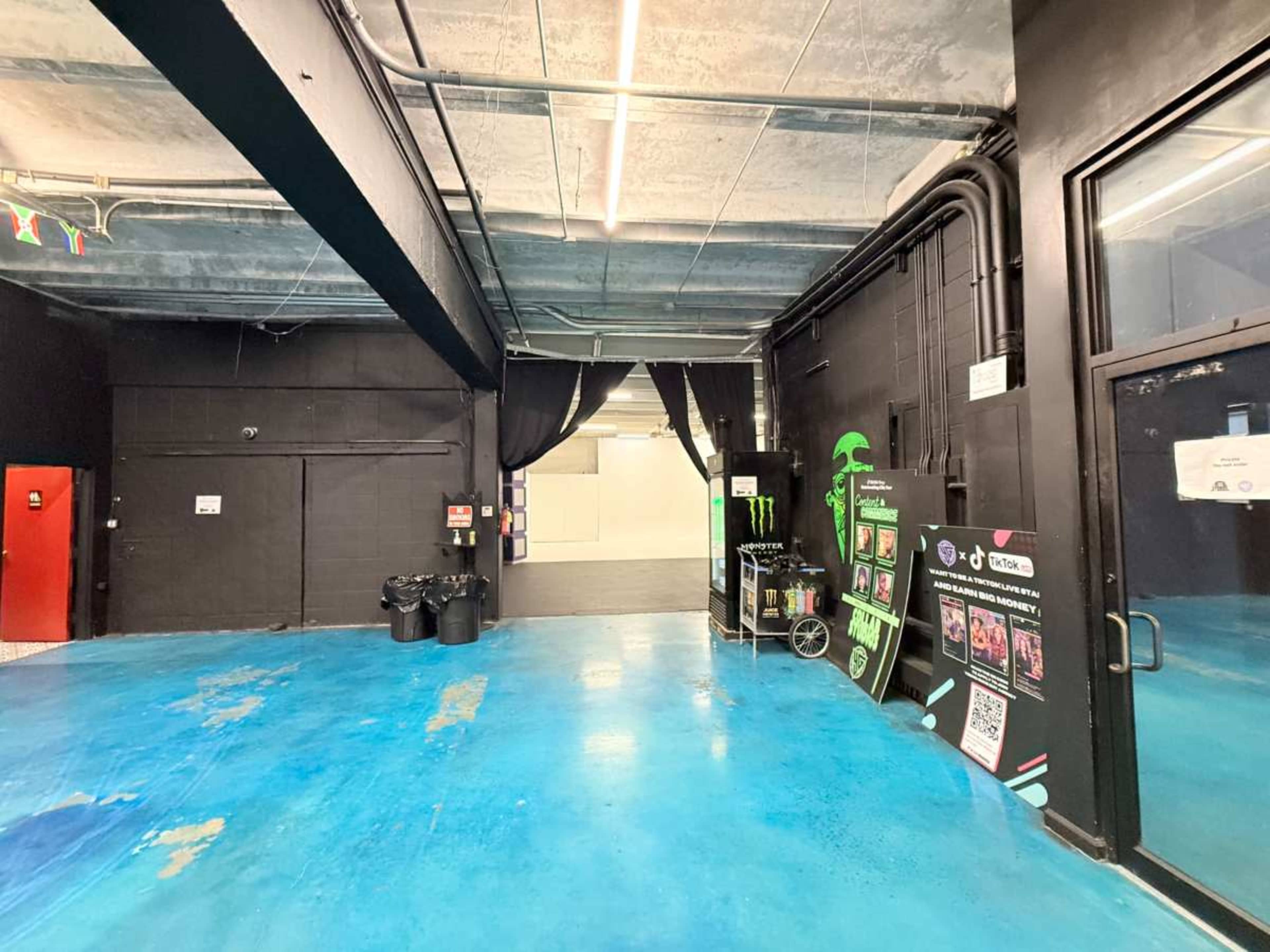 The image shows a large, empty room with a blue floor, black walls, and a doorway leading to another area, along with a display featuring Monster Energy drinks.