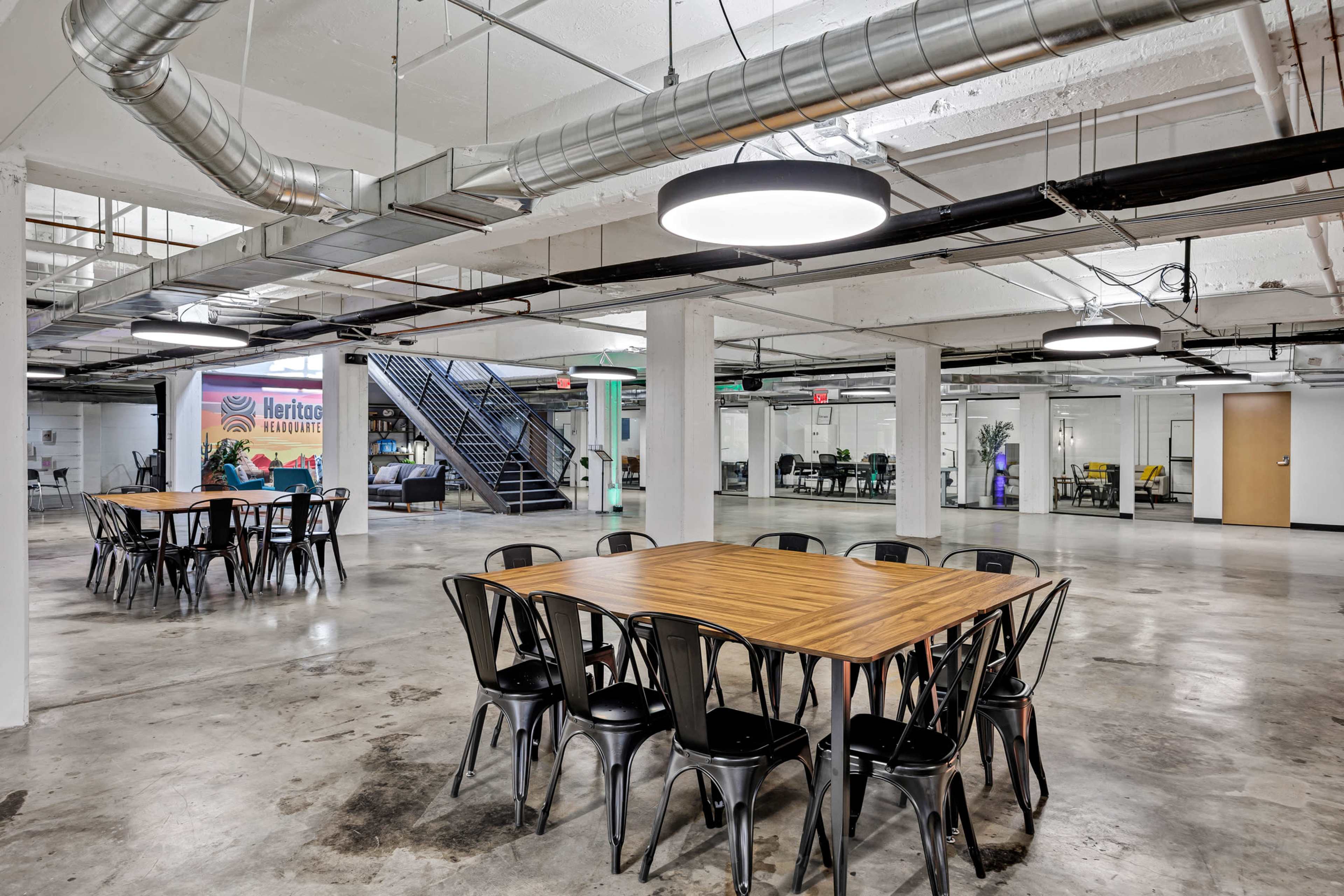 The image shows a spacious, modern office interior with exposed ceilings, multiple tables arranged for meetings, and glass-walled conference rooms in the background.