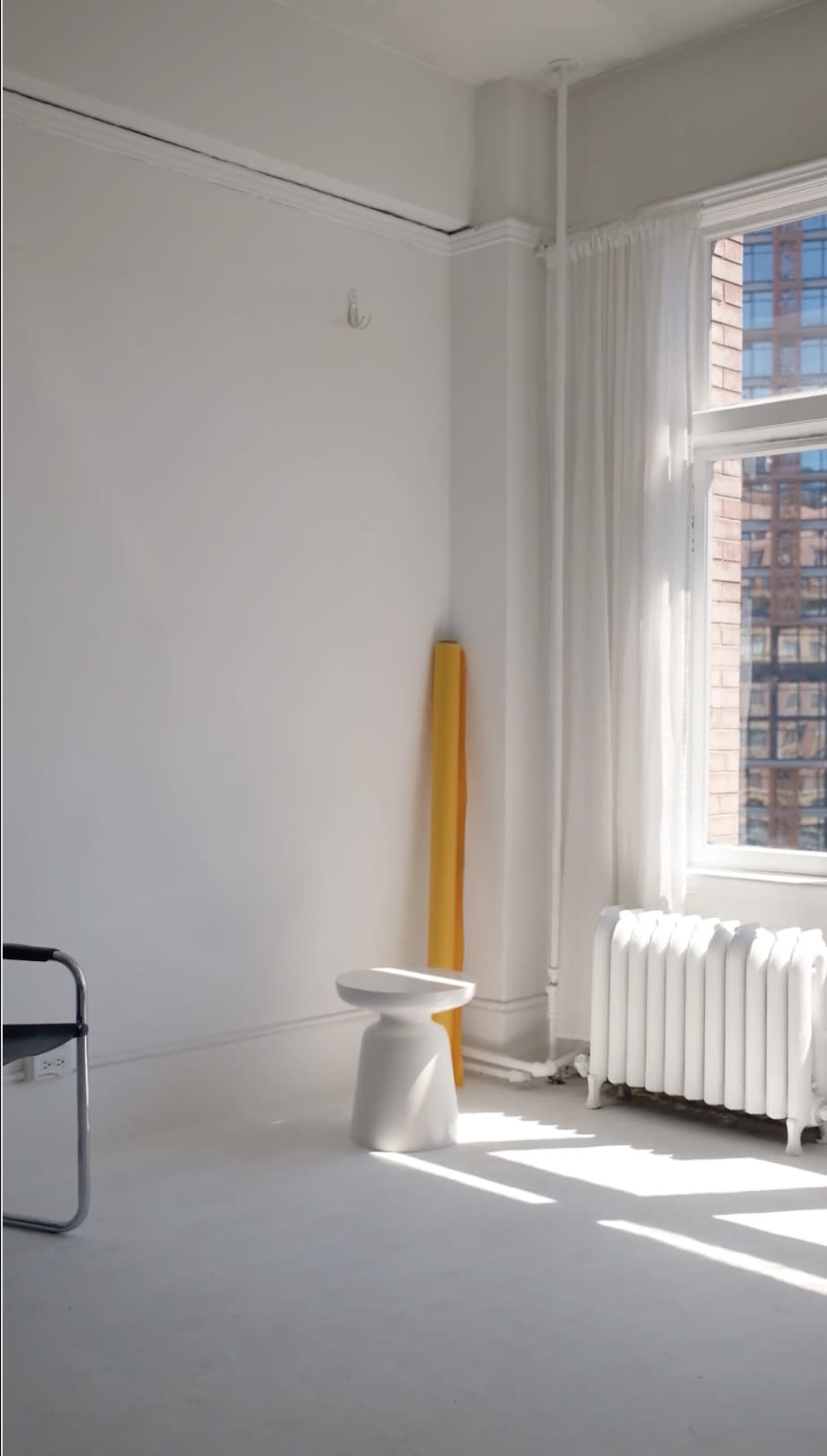 The image shows a minimalist interior with a white wall, a stool, a yellow pole, and natural light streaming through a large window.