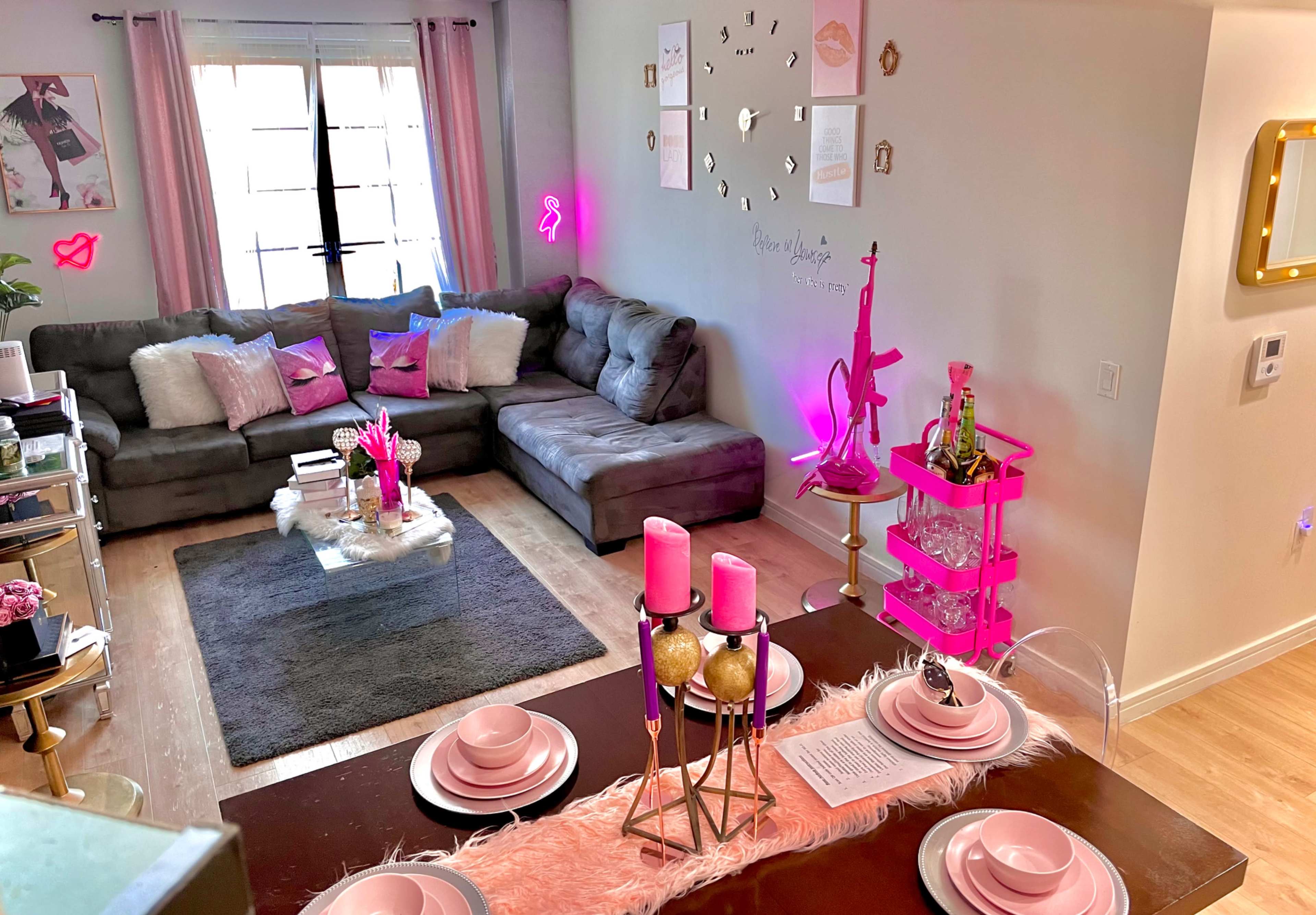 A modern living space with a gray sectional sofa, a dining table set with pink decor and candles, and decorative elements like a pink cart and wall art.