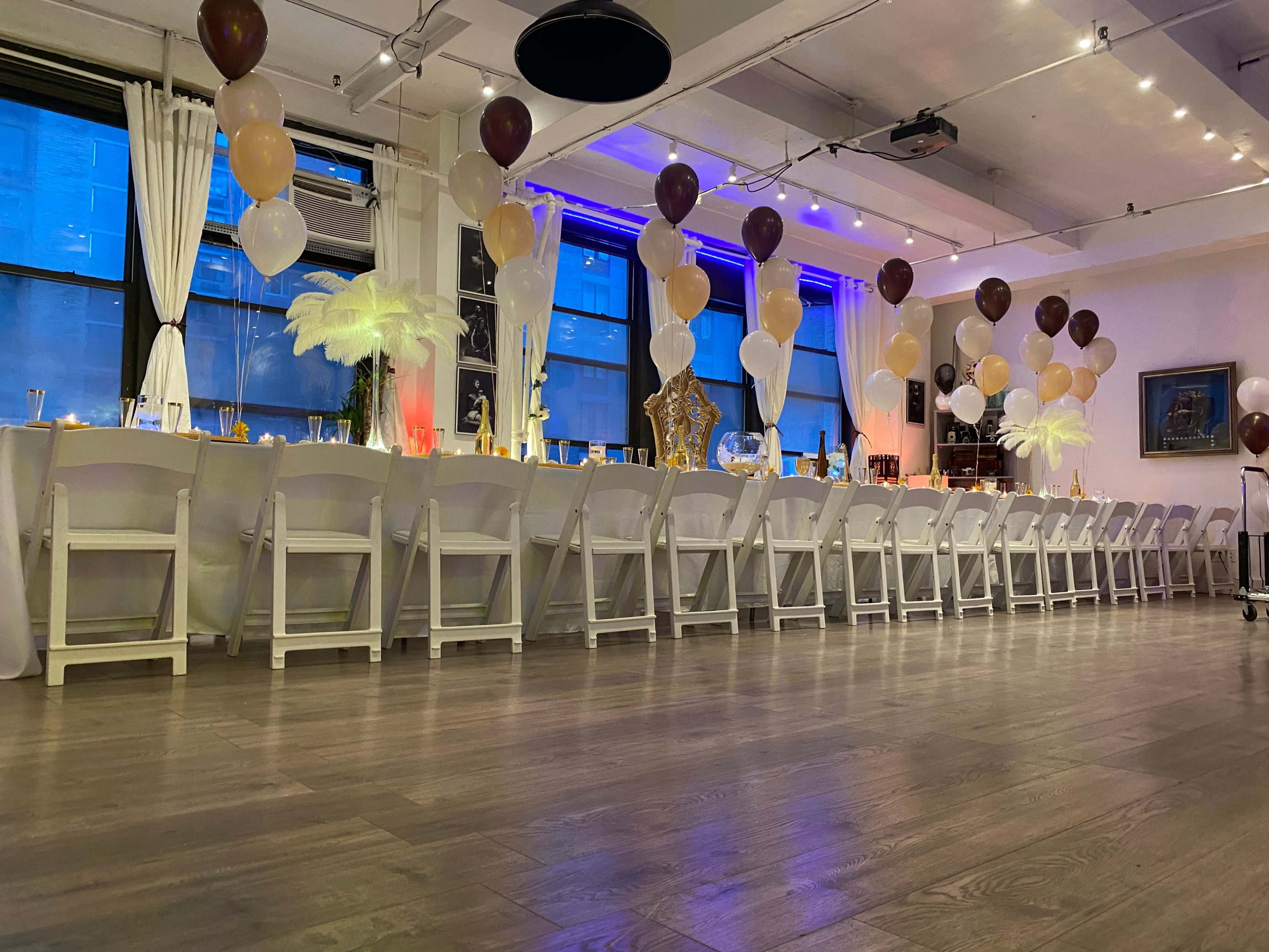 A banquet setup in a bright room, featuring a long table adorned with white tablecloths, gold and brown balloons, and decorative elements.