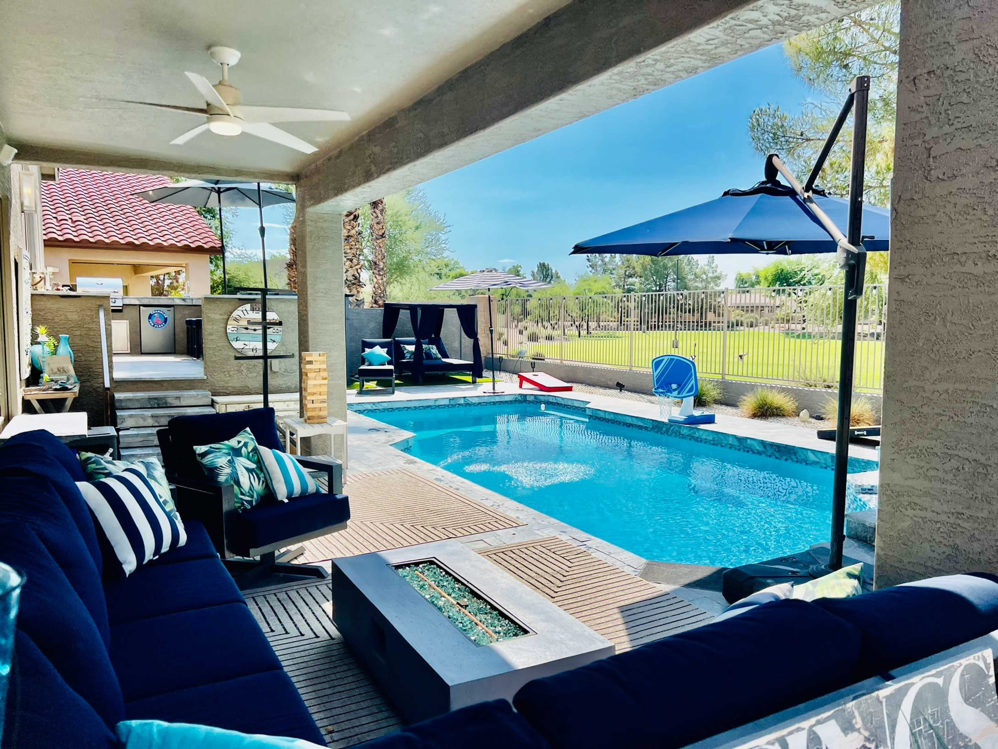 A modern patio area features a swimming pool, lounge seating, and umbrellas, surrounded by a manicured lawn and palm trees.