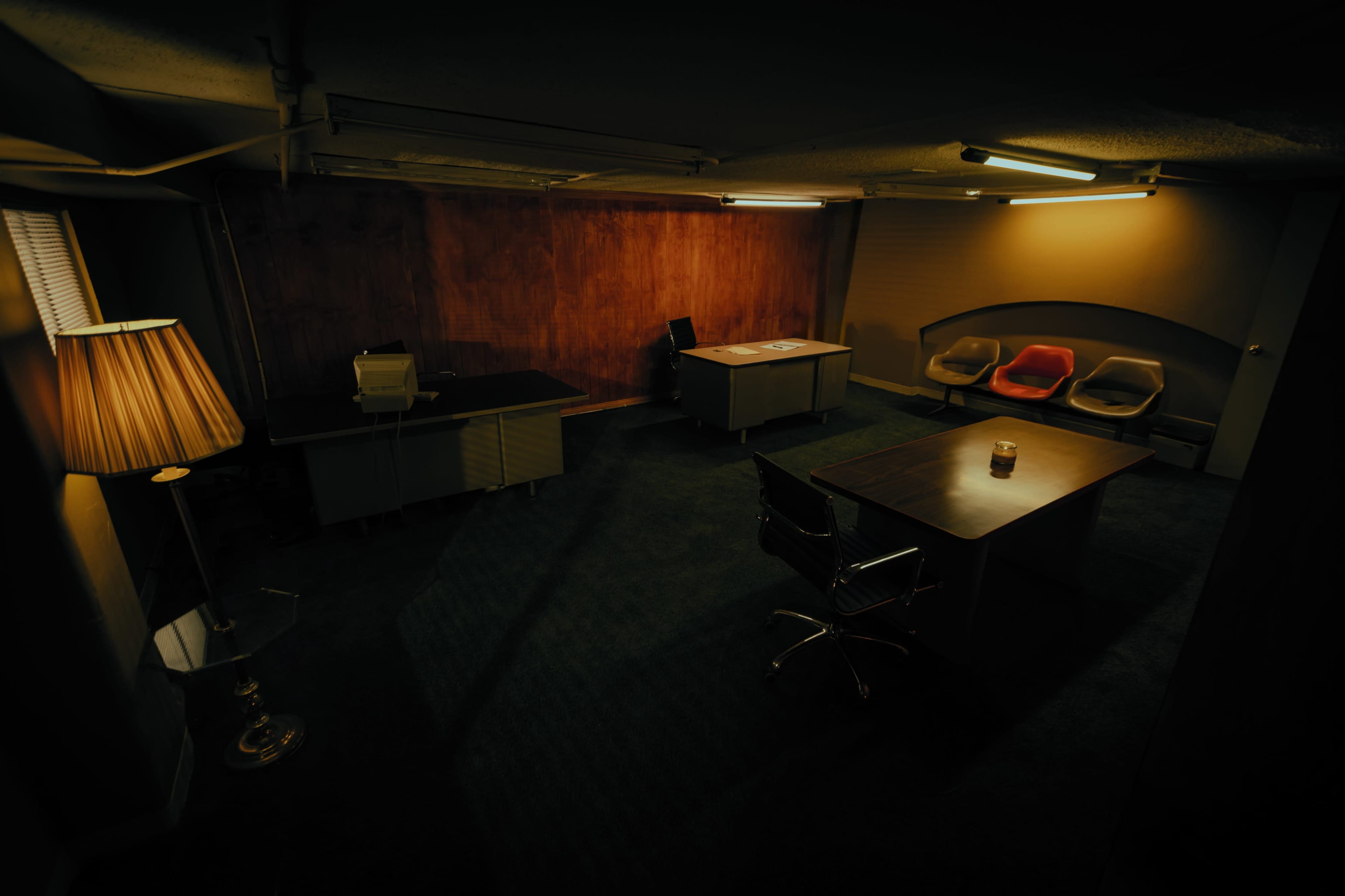 A dimly lit office space featuring two desks, a lamp, and a set of chairs.