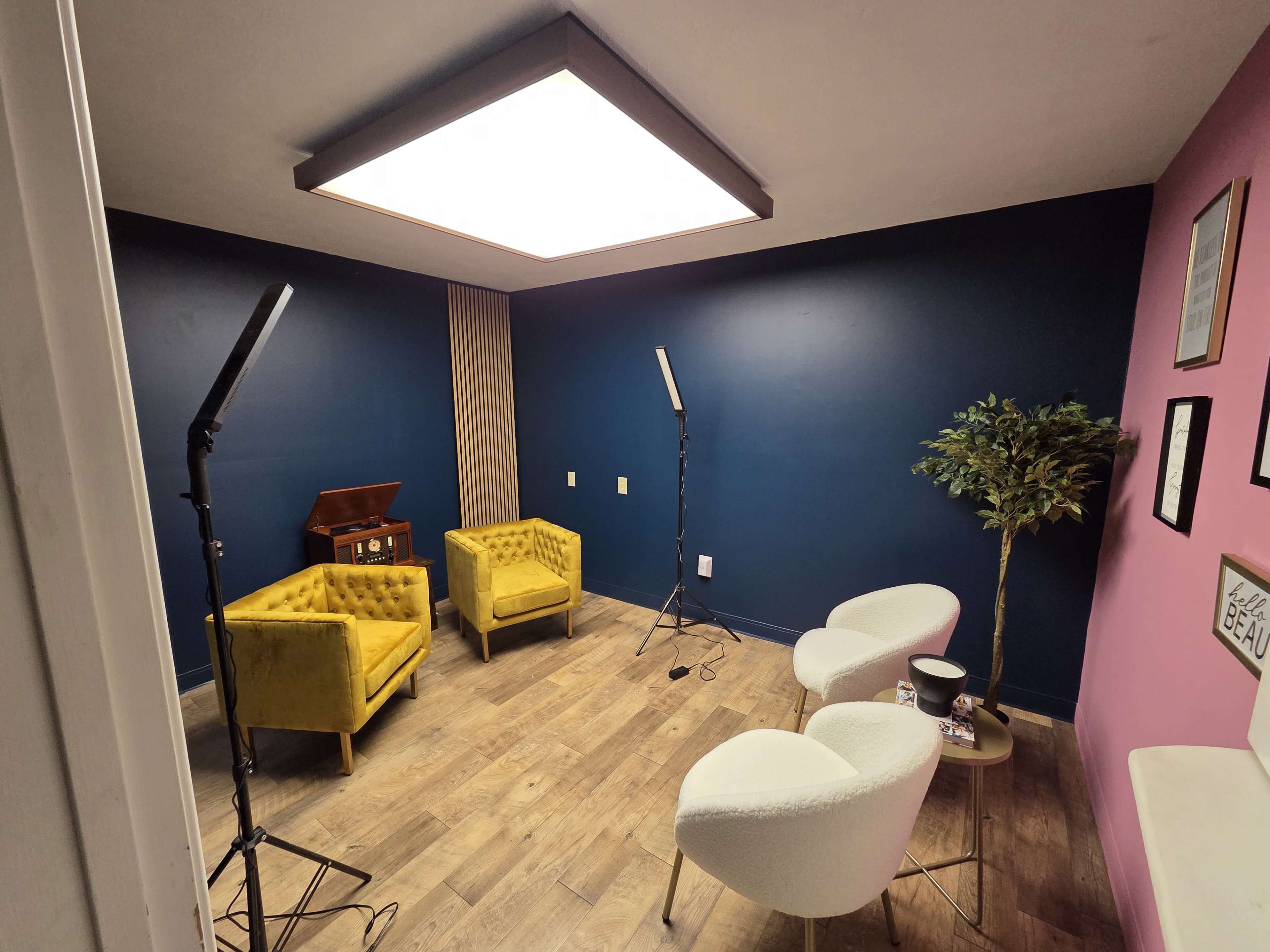 The image shows a small room with blue and pink walls, featuring two yellow chairs, a white chair, a small table, and lighting equipment set up for filming or photography.