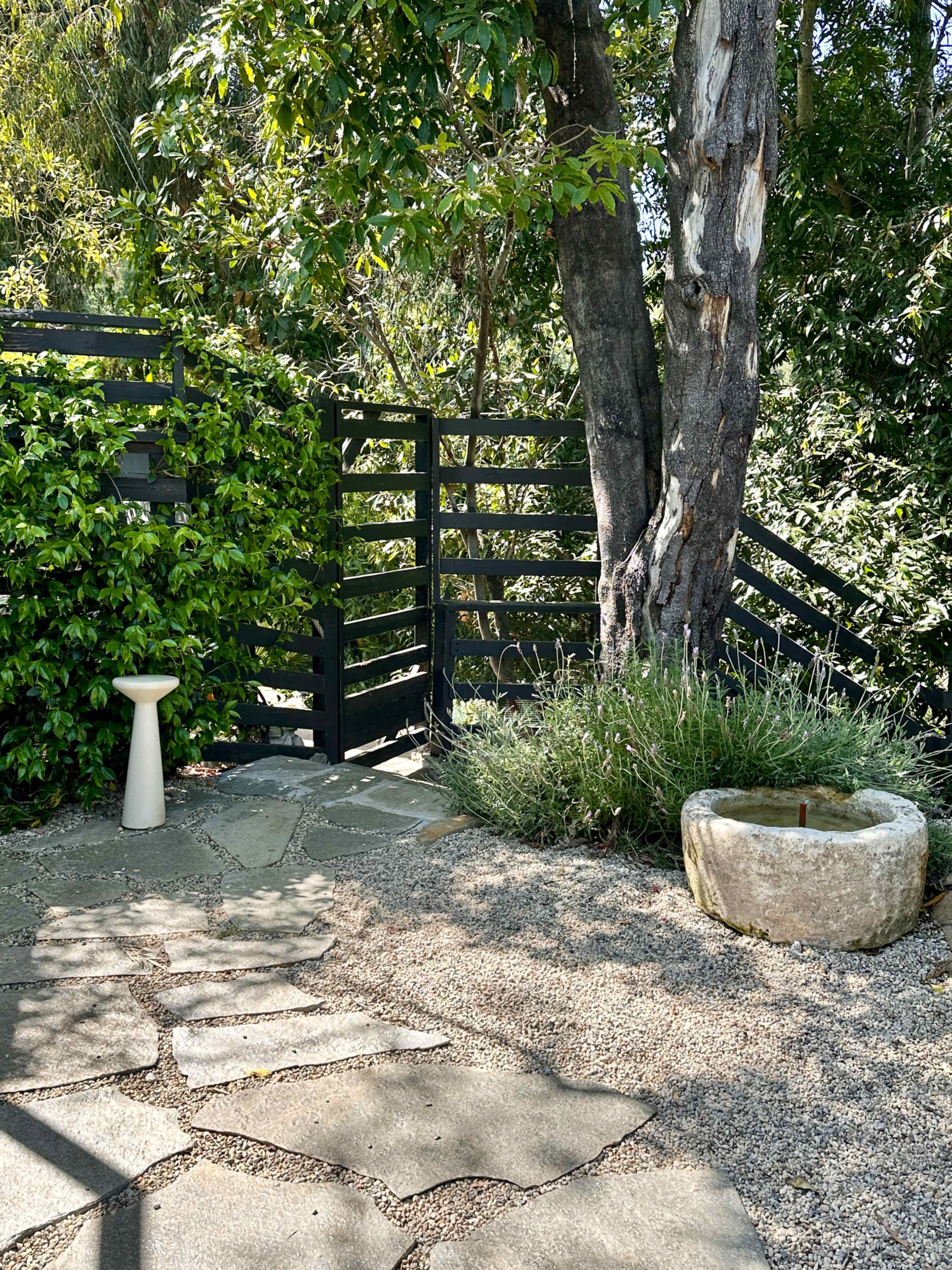 A stone pathway leads to a fenced garden area with a tree, a birdbath, and a circular stone feature.