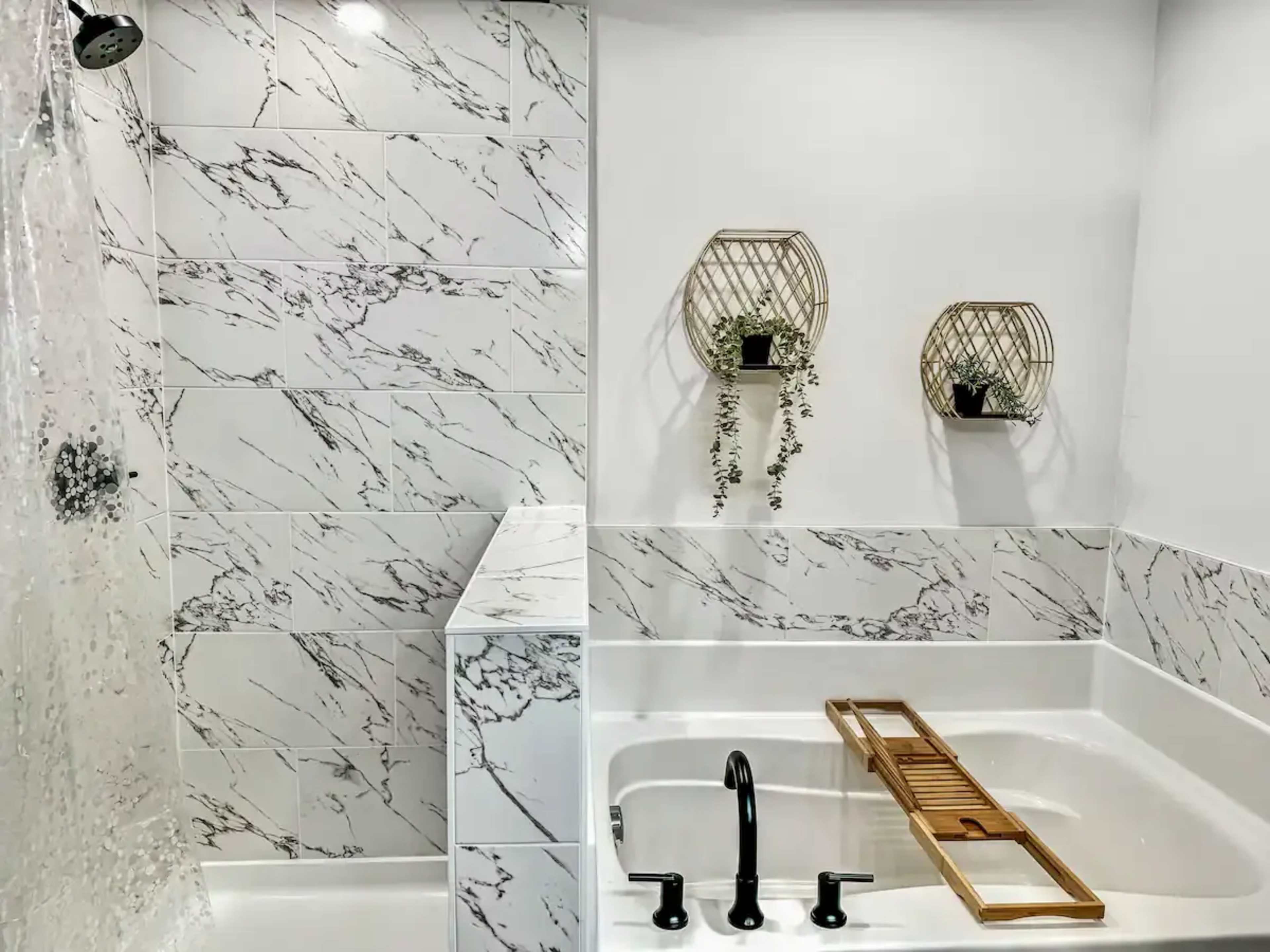 The image shows a modern bathroom featuring a marble-patterned tiled shower, a white bathtub with a wooden tub tray, and decorative wall planters.
