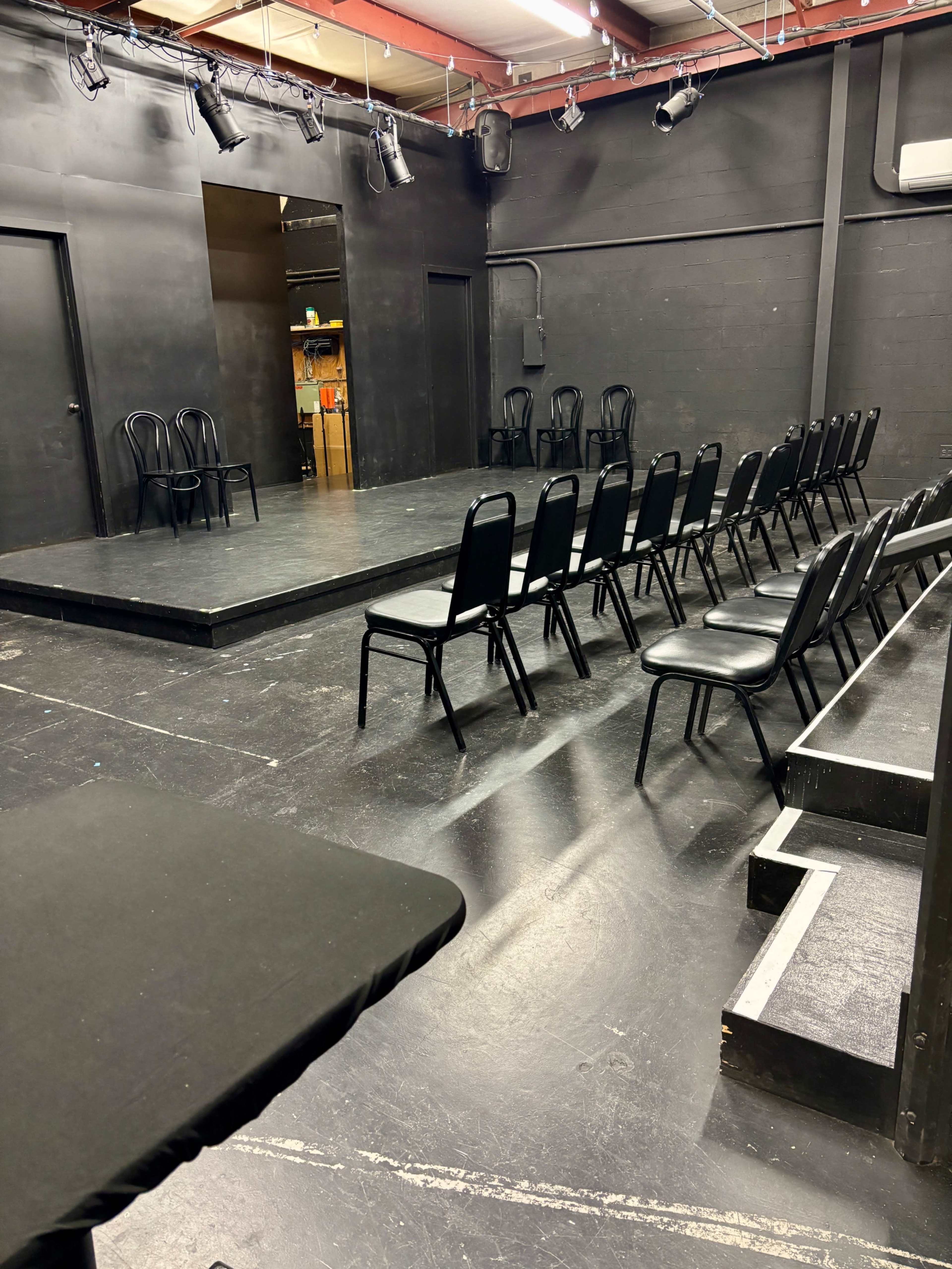 The image shows a small, black-walled theater stage with a raised platform surrounded by rows of black chairs facing the stage.