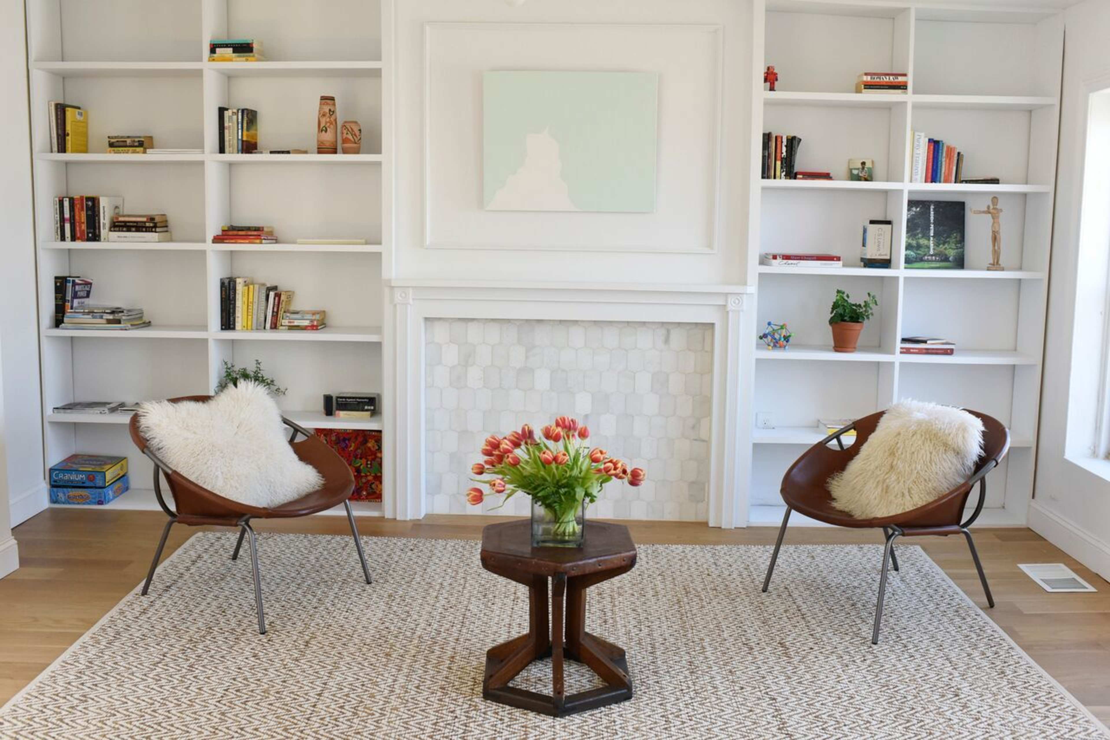 A modern living room features two brown chairs, a round table with a vase of tulips, and white bookshelves filled with books and decorative items.