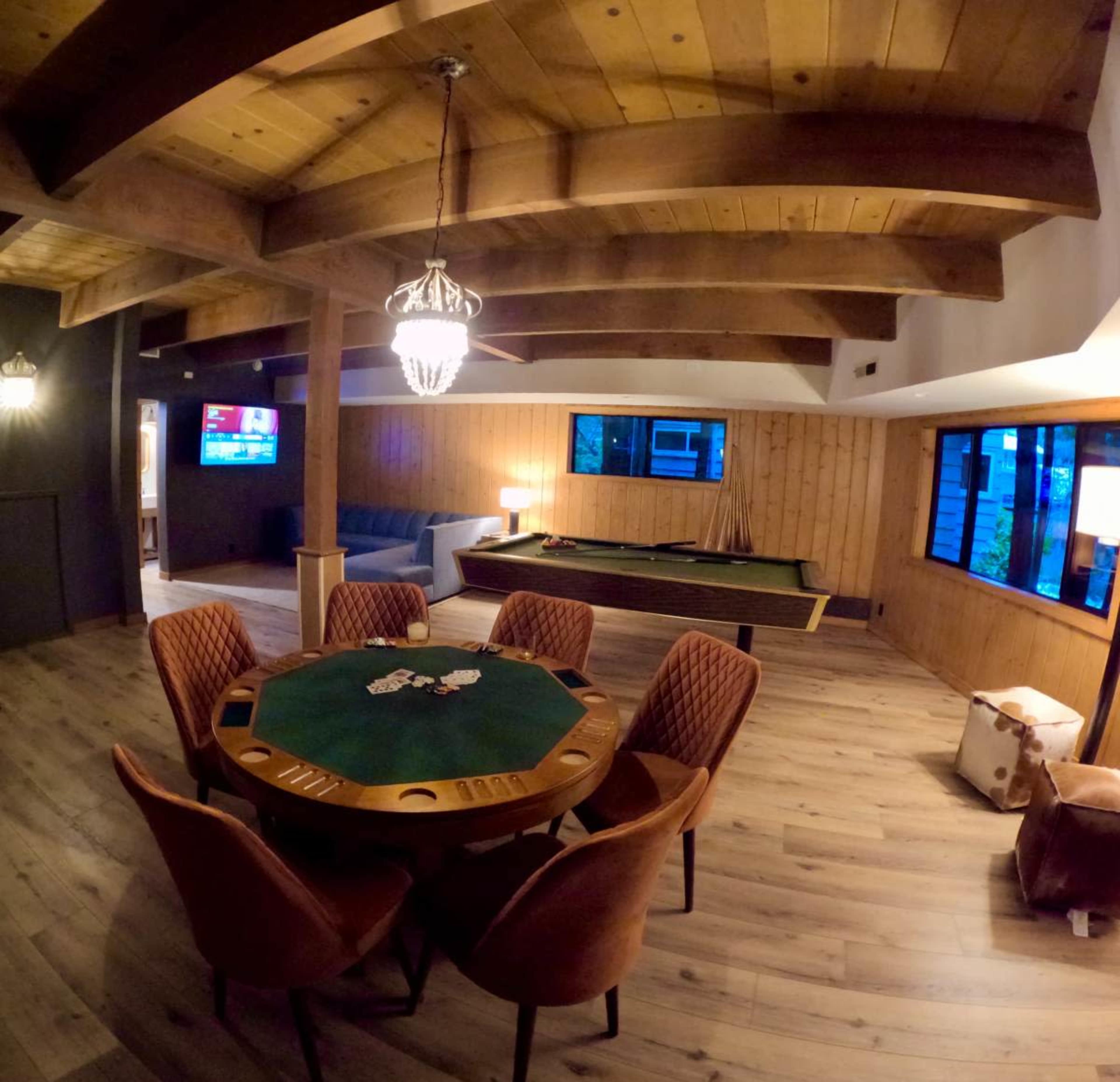 The image shows a spacious room with wooden beams, a poker table surrounded by chairs, a pool table, and large windows allowing natural light.