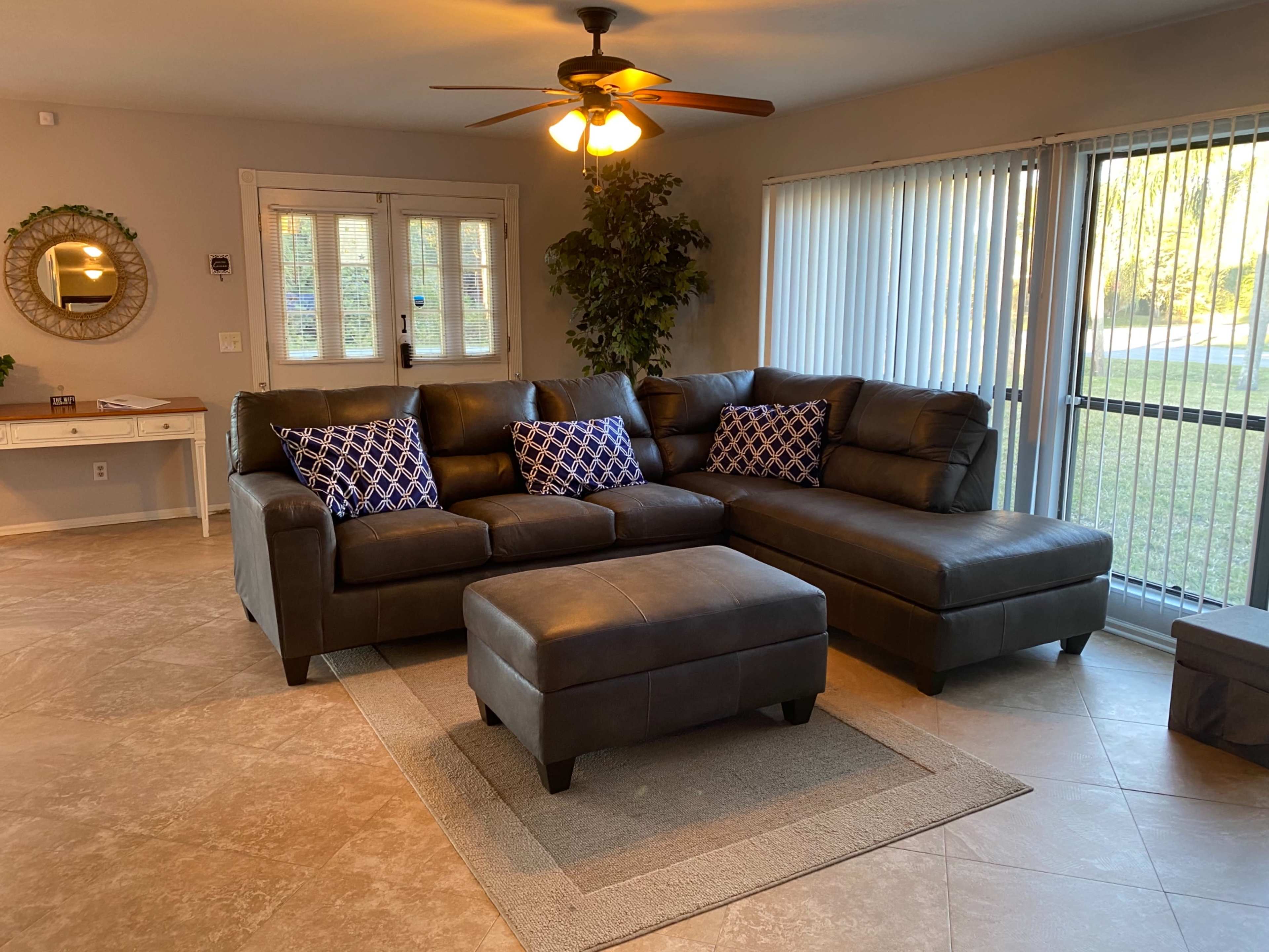 A living room with a sectional leather sofa, a large ottoman, and a sliding glass door, illuminated by ceiling lights.