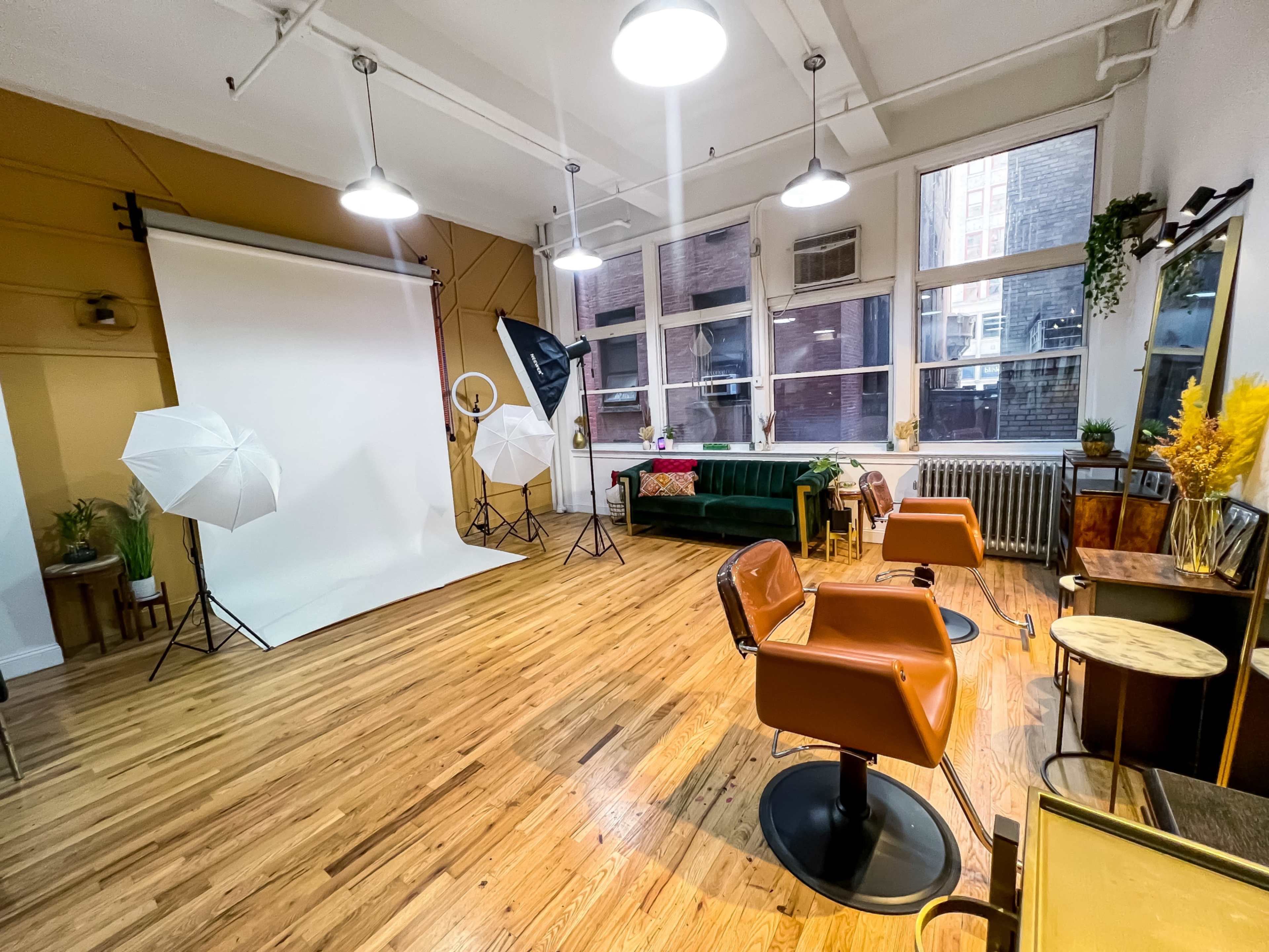 A spacious studio setup with a white backdrop, two softbox lights, a green couch, and several salon chairs on a wooden floor.