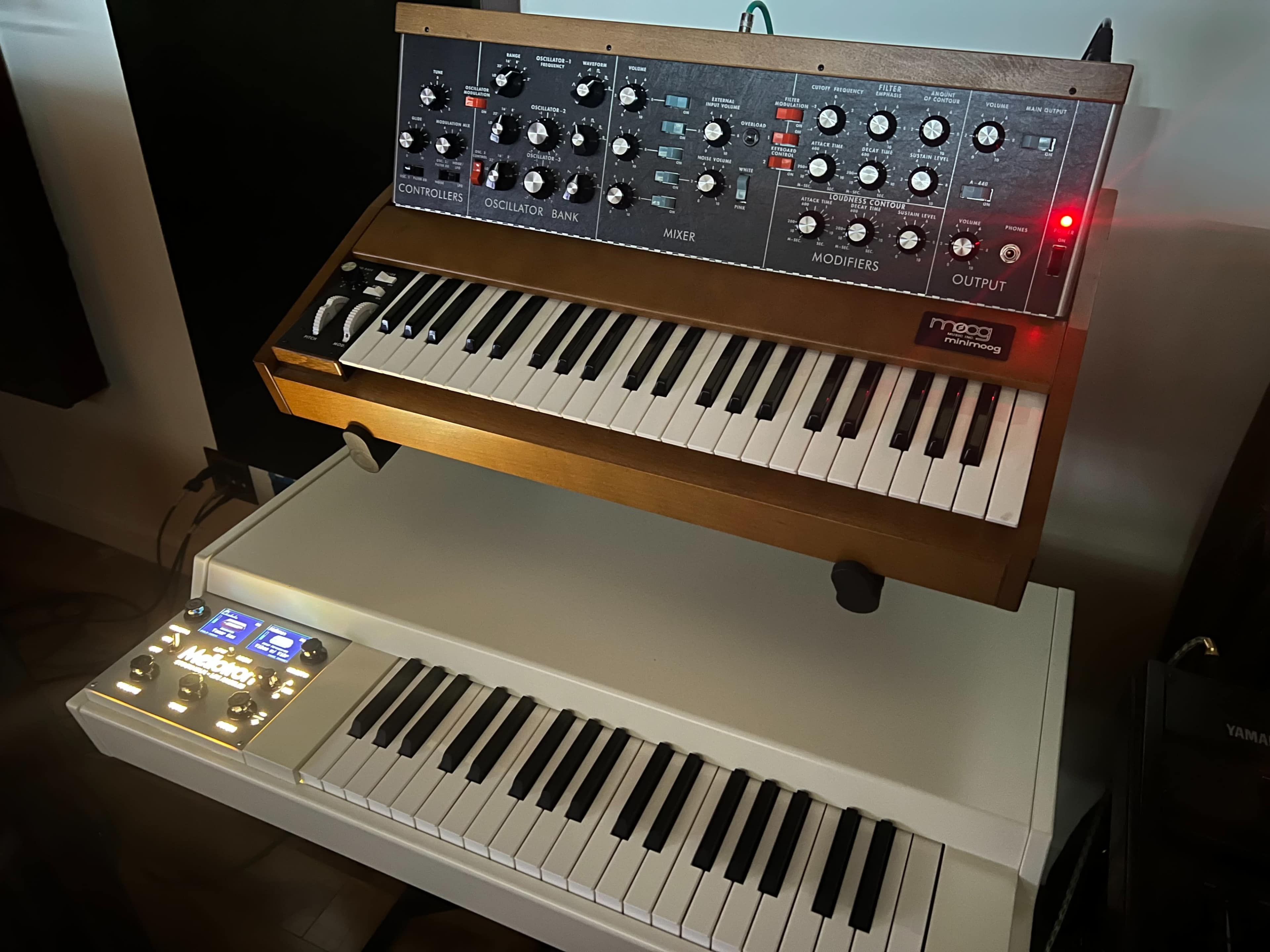 The image shows a Moog synthesizer with a wood finish mounted above a white keyboard, both positioned on a surface in a well-lit room.