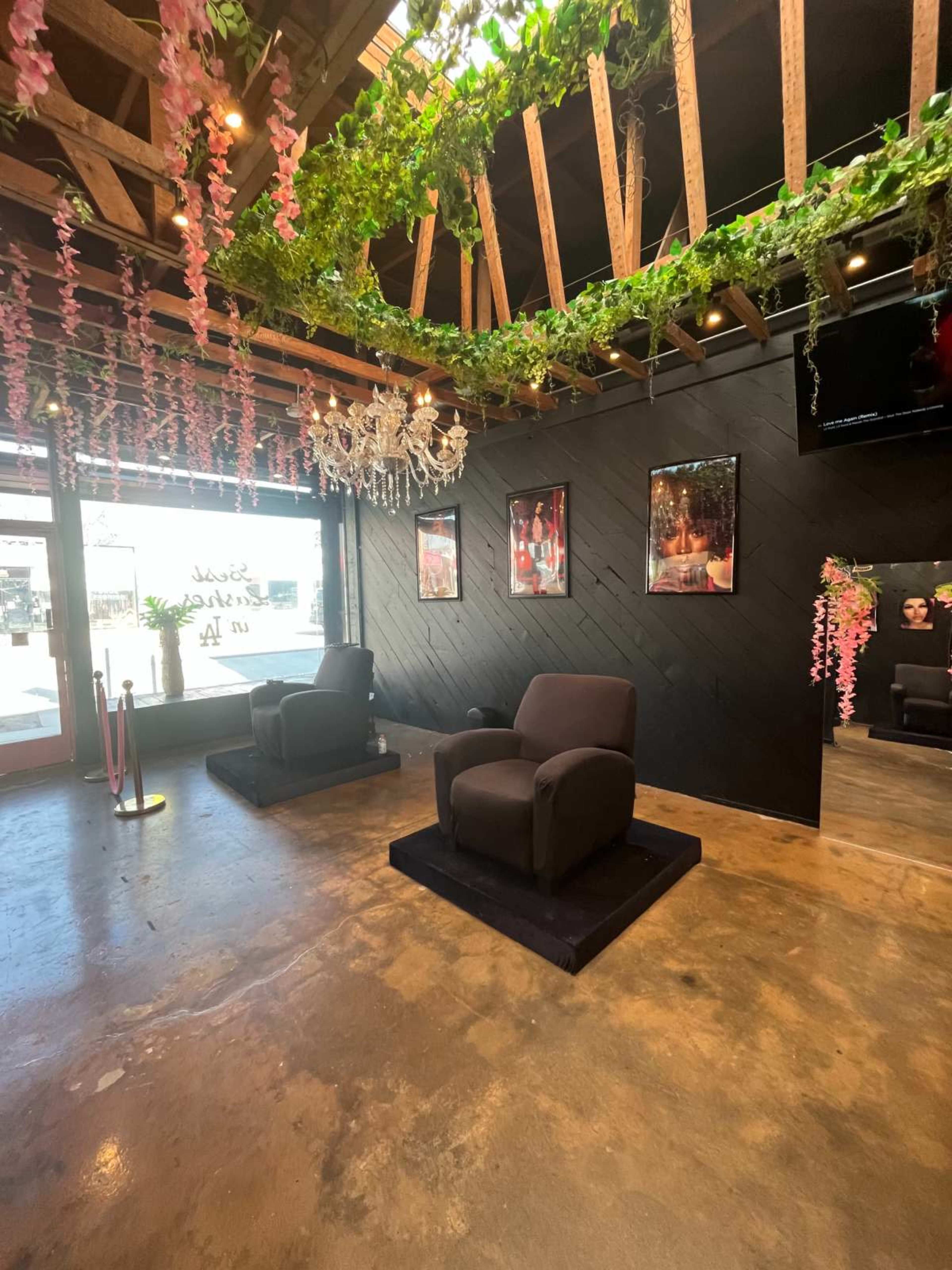 The interior of a salon features two dark chairs on a black platform, surrounded by greenery and pink floral decorations hanging from the ceiling.