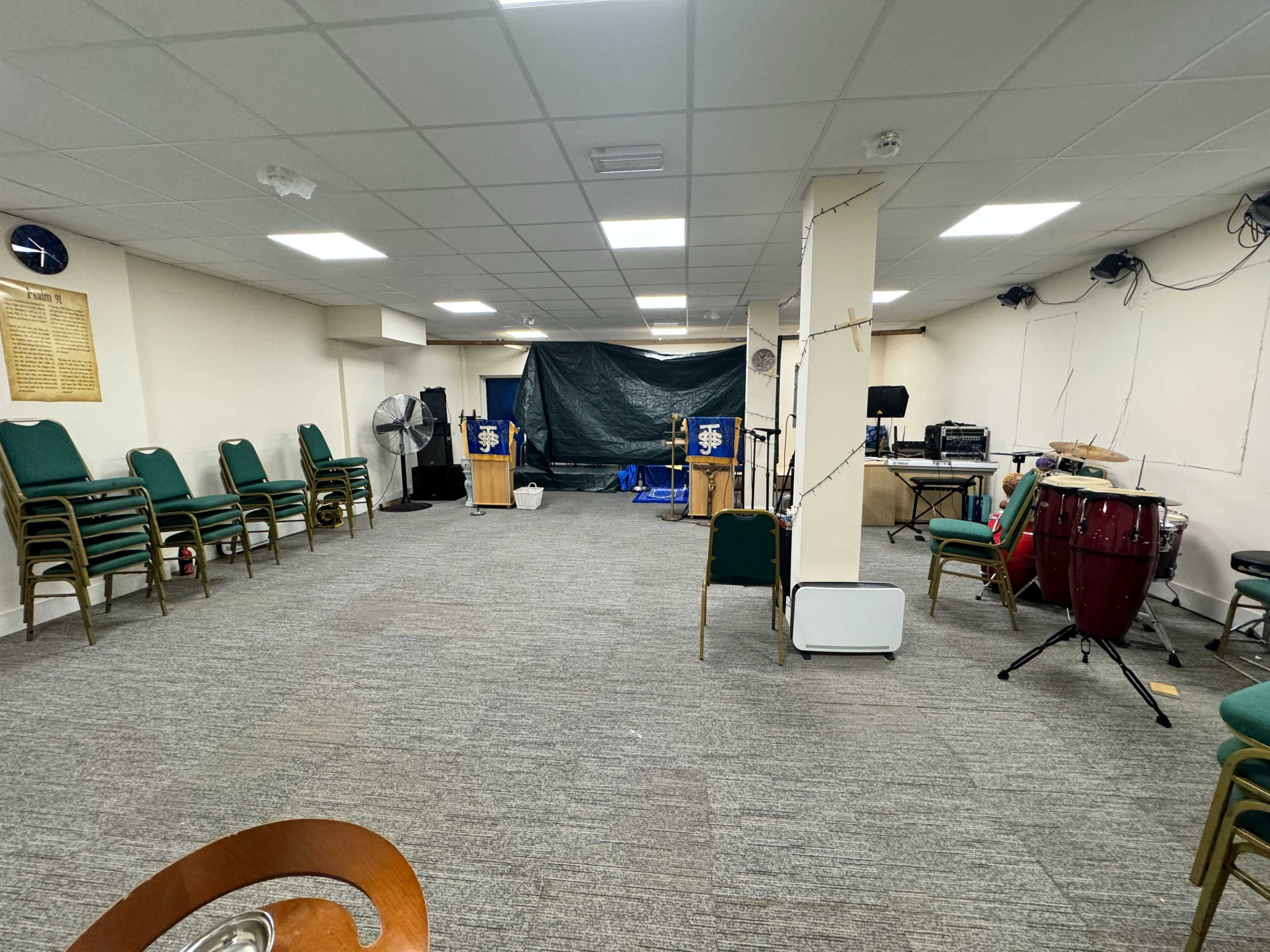 The image shows an empty indoor space with green chairs stacked against the wall, a podium at the front, and a percussion instrument on the side.