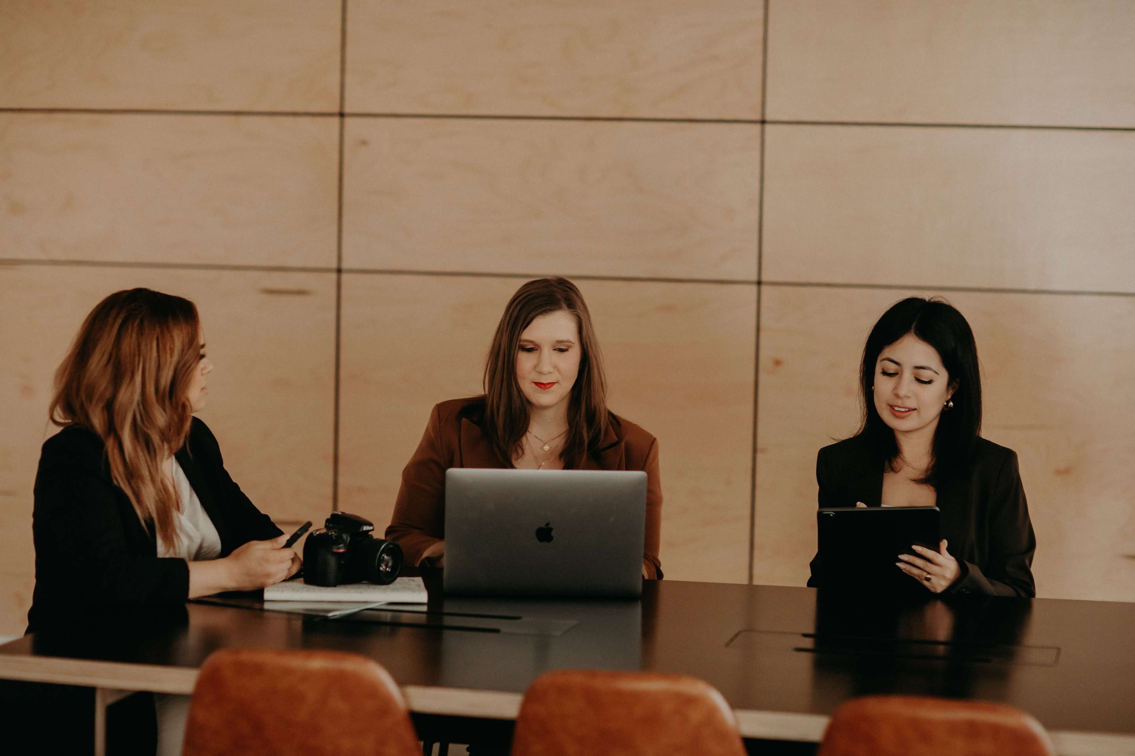 Three women are seated at a conference table, each engaged with technology, including a laptop and camera, in a modern office environment.
