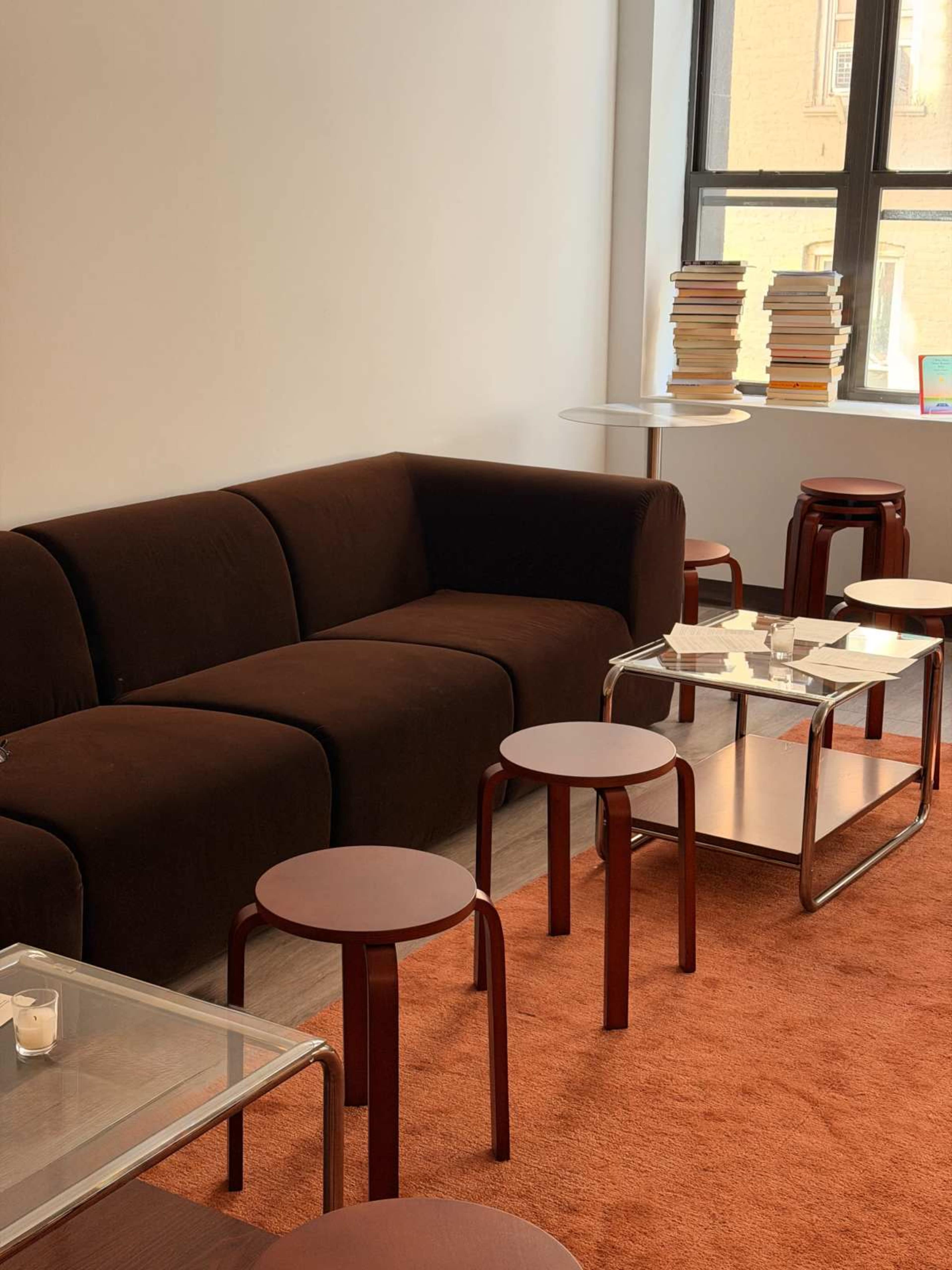 The image shows a modern lounge area featuring a brown couch, multiple small round tables, and a stack of books near a window.