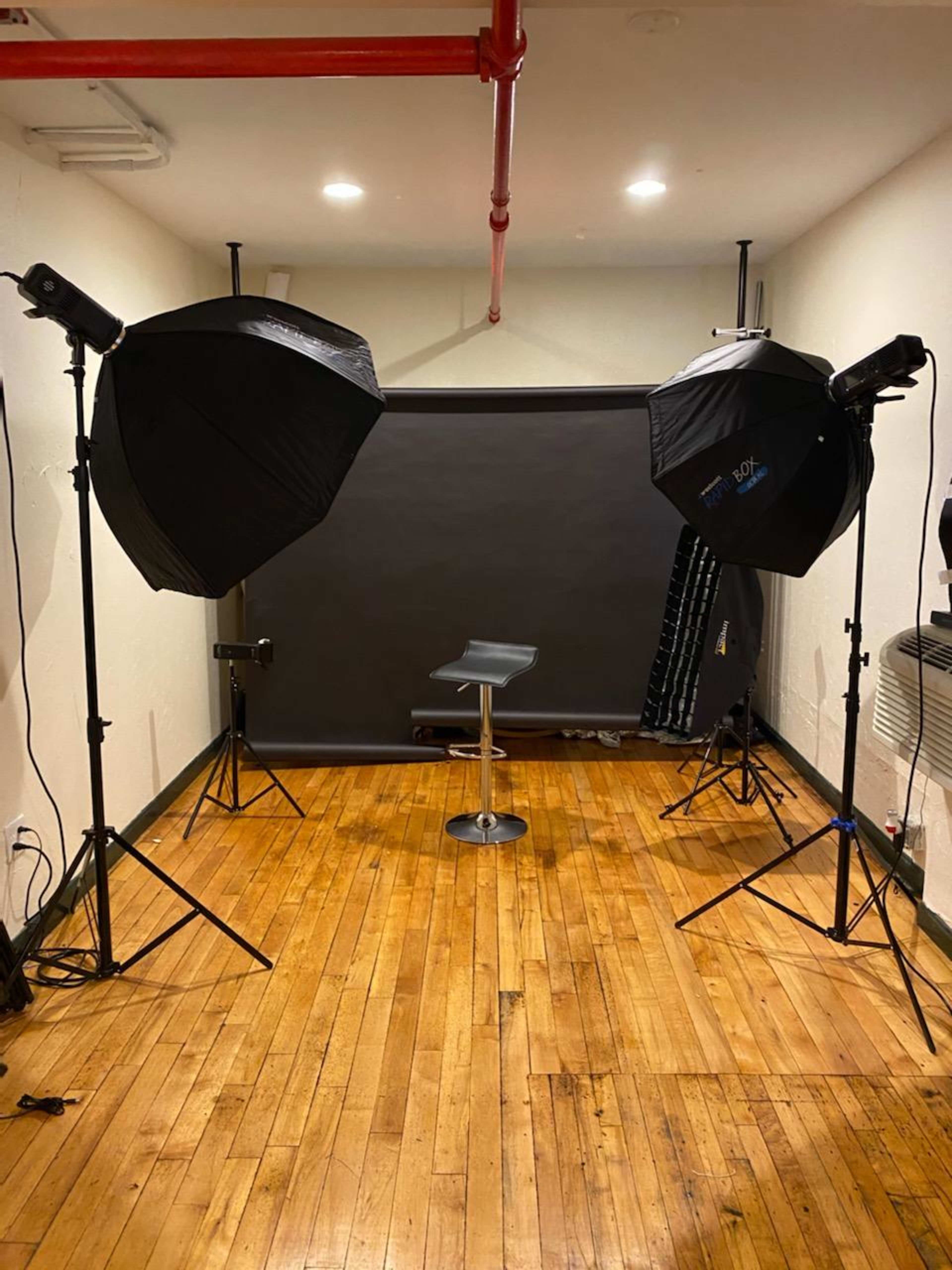 The image shows a photography studio setup with two softbox lights positioned on either side of a black backdrop and a stool in the center.