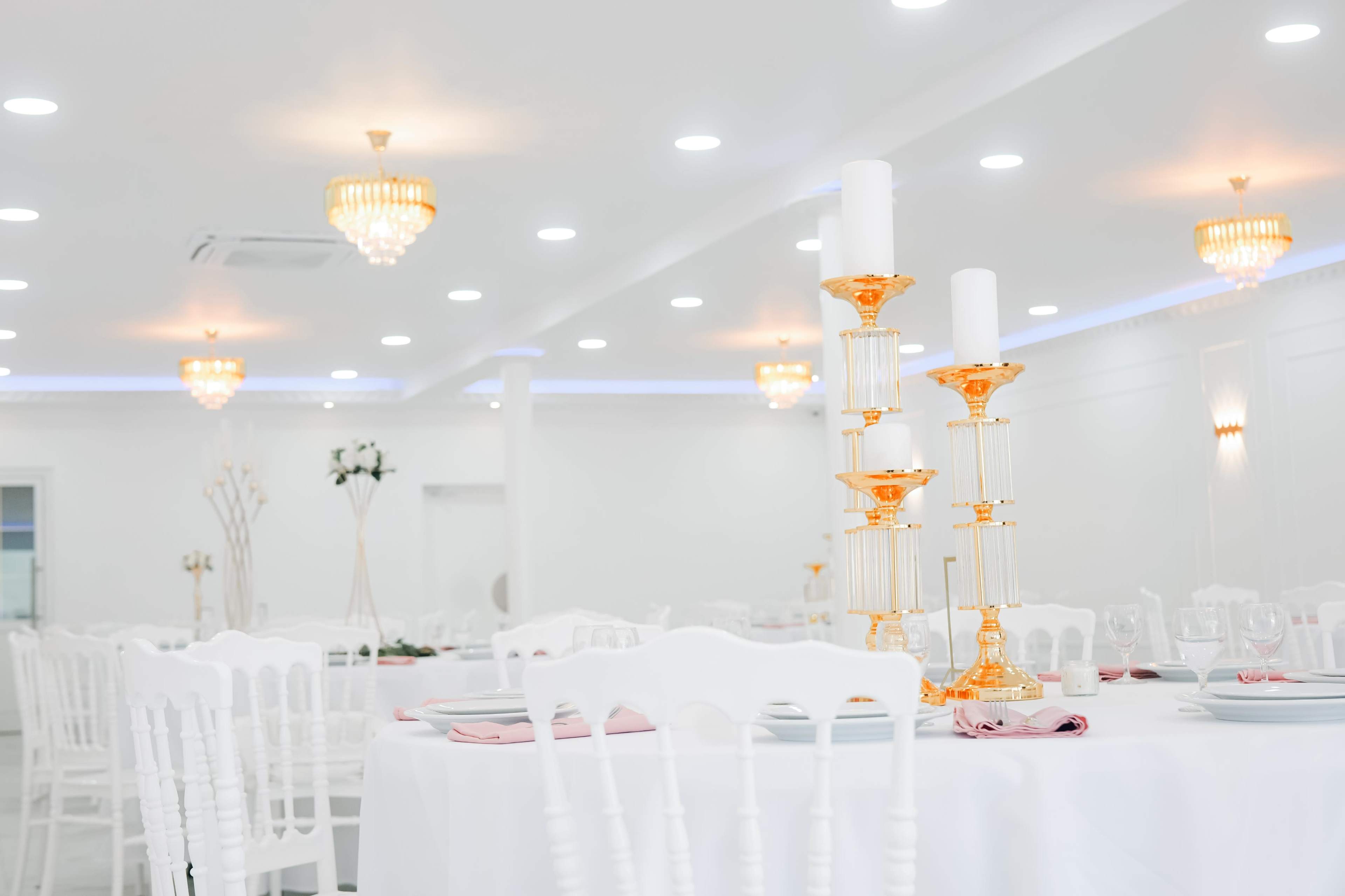 The image shows a white banquet hall with elegant gold-accented decor, featuring white chairs and tables set for an event.