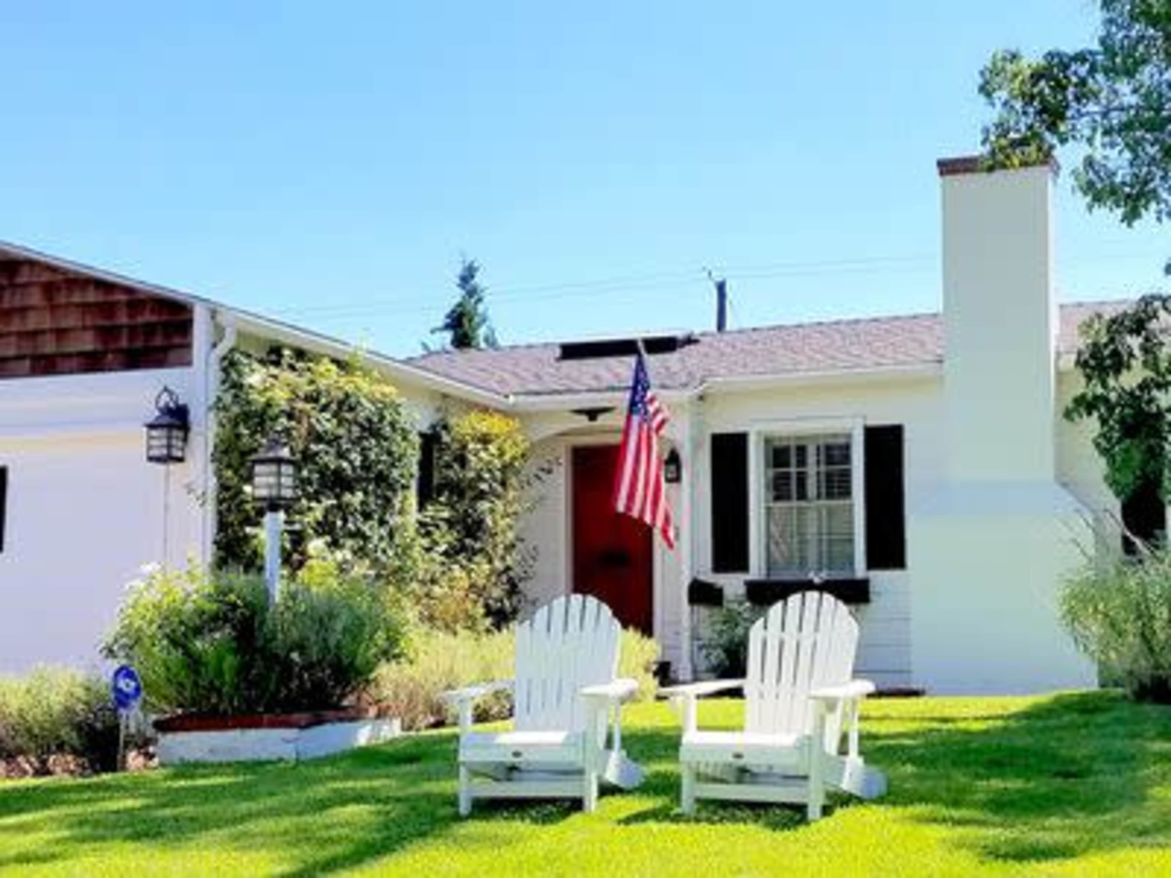 A white house with black shutters and an American flag stands in front of a well-manicured lawn featuring two white adirondack chairs.