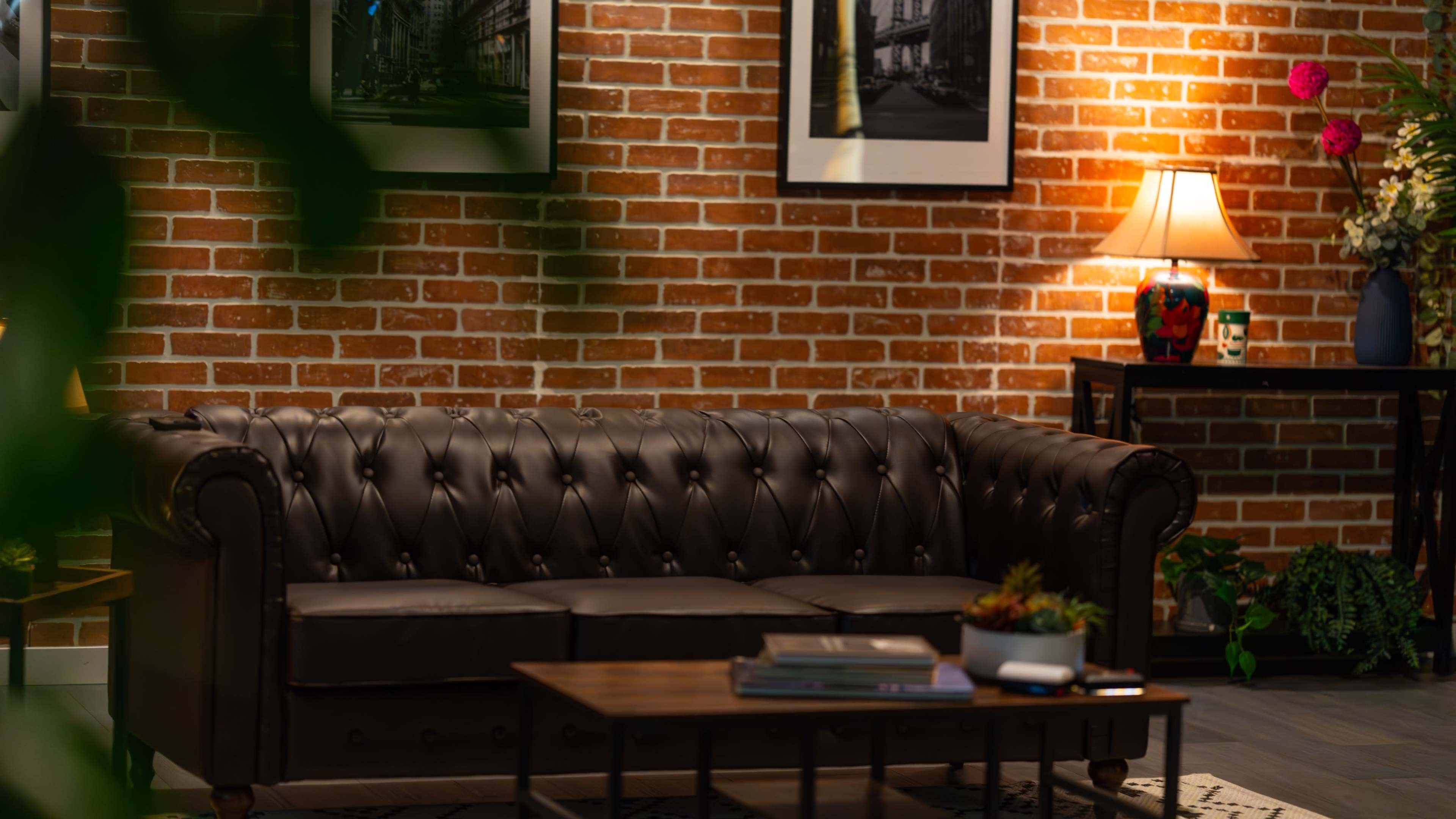The image shows a leather chesterfield sofa in front of a brick wall, accompanied by a wooden coffee table and a lamp on a side table.