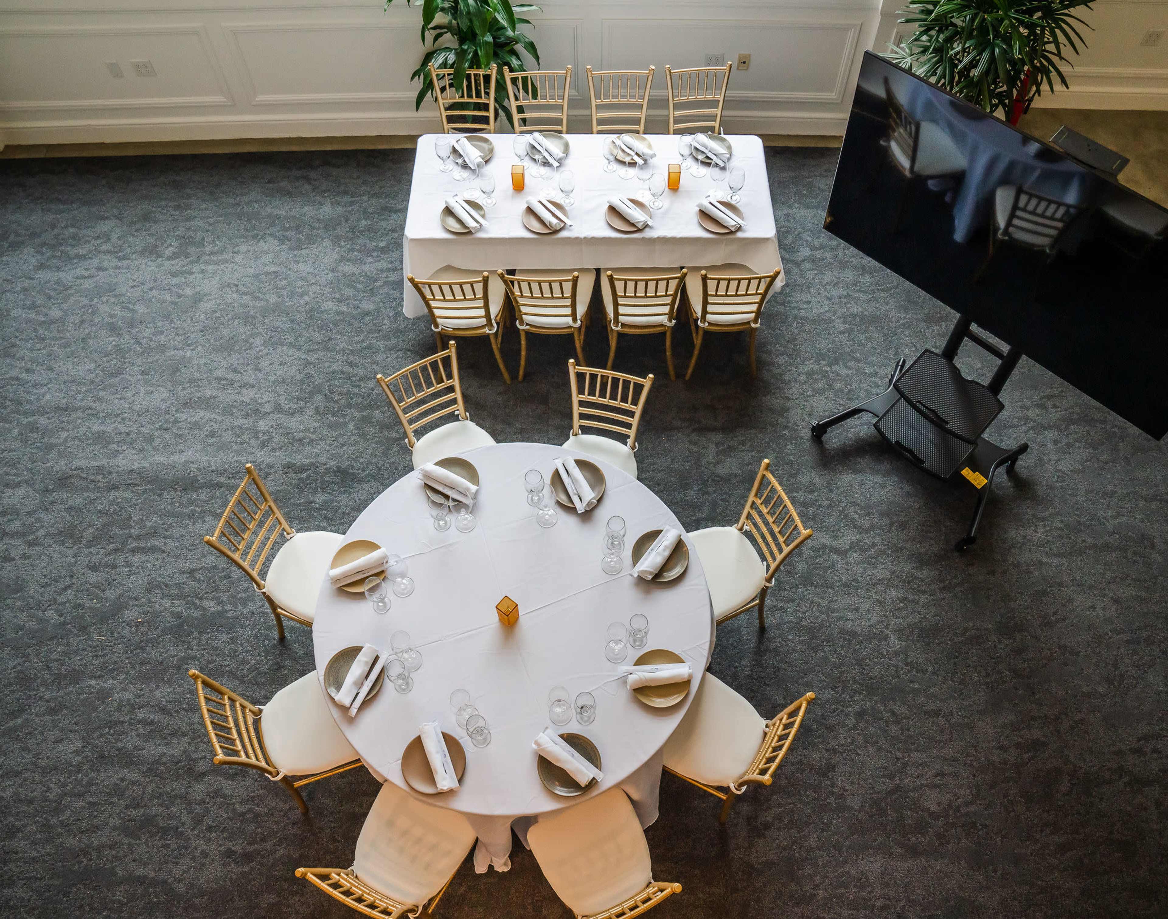 A spacious dining area with two tables set with white tablecloths, elegant place settings, and surrounded by gold-colored chairs, alongside a television screen on a mobile stand.