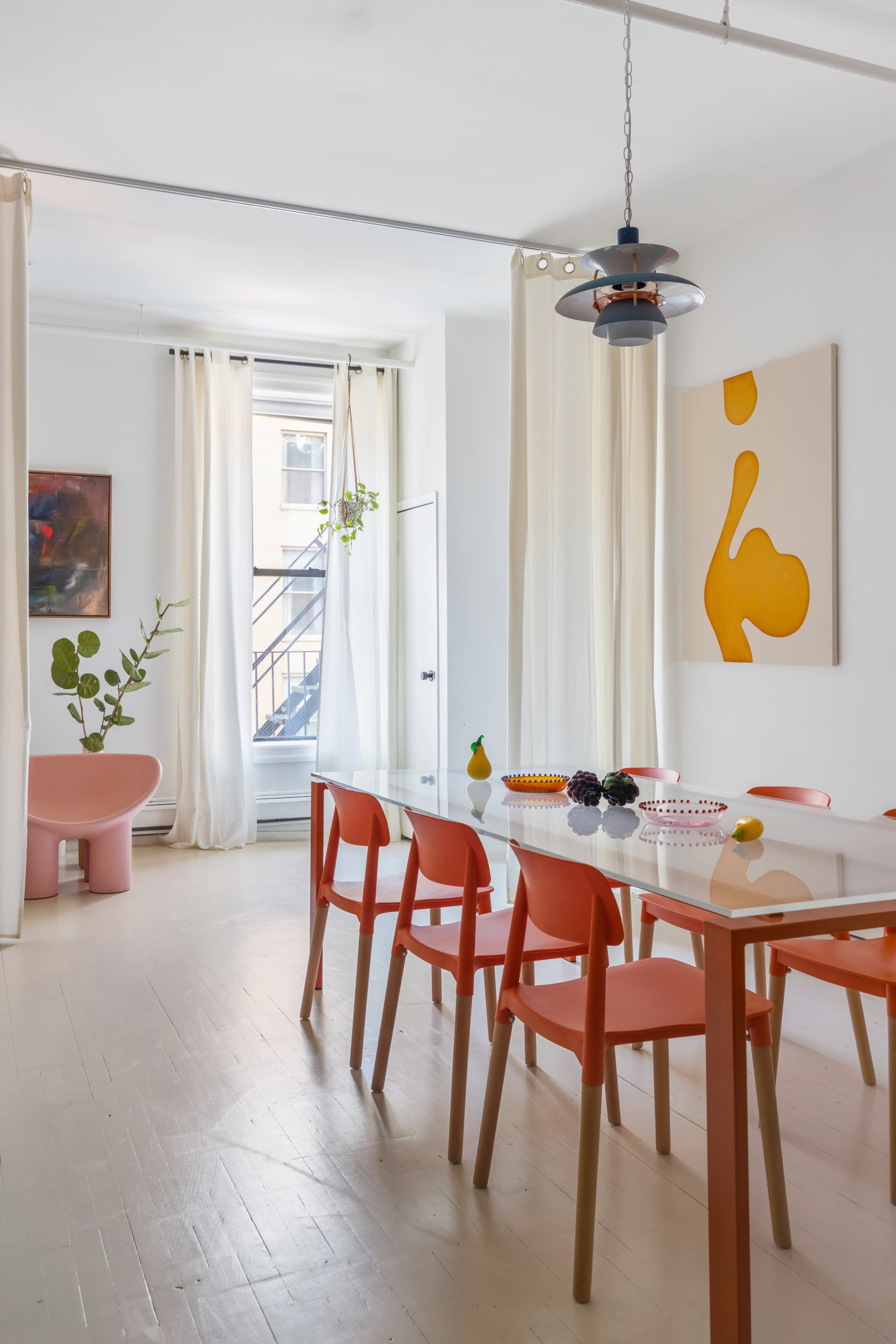 A bright dining area features a white table surrounded by orange chairs, with curtains, artwork, and decorative objects enhancing the modern aesthetic.