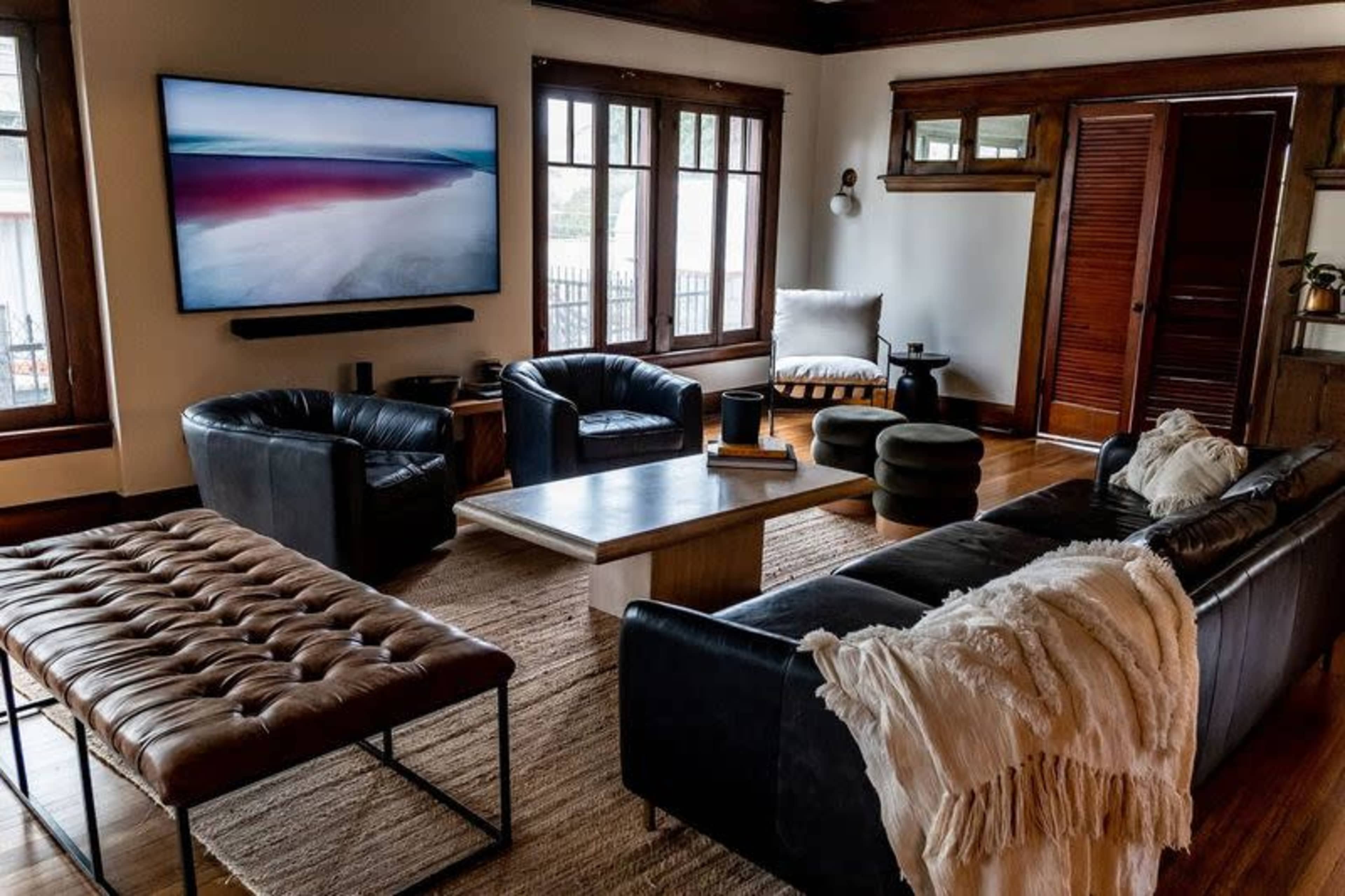 The living room features a large TV mounted on the wall, a dark leather sofa, two leather armchairs, a wooden coffee table, and a textured area rug.