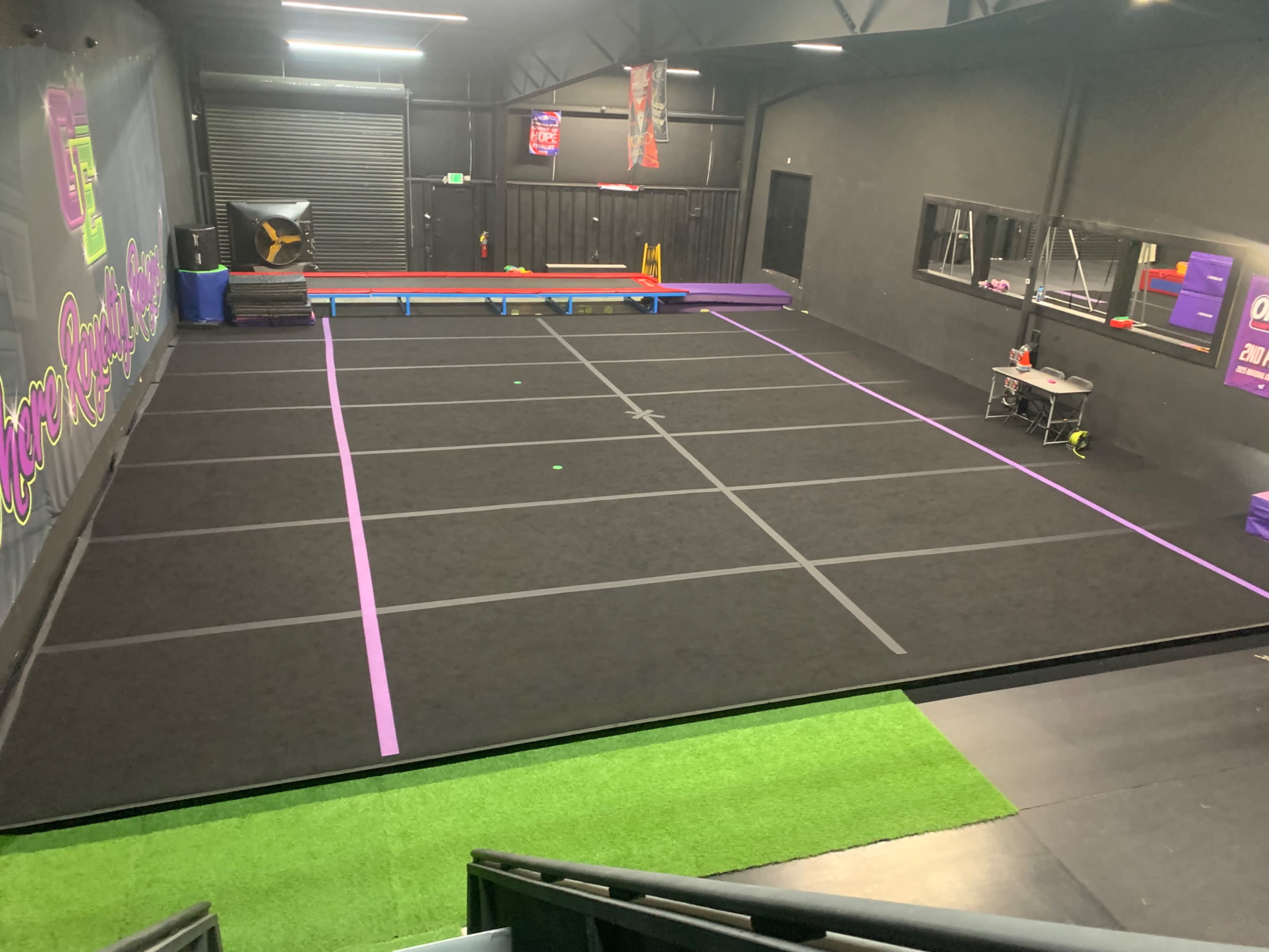 The image shows an indoor gymnasium with black flooring, marked by purple lines, and equipped with various training apparatuses along the walls.