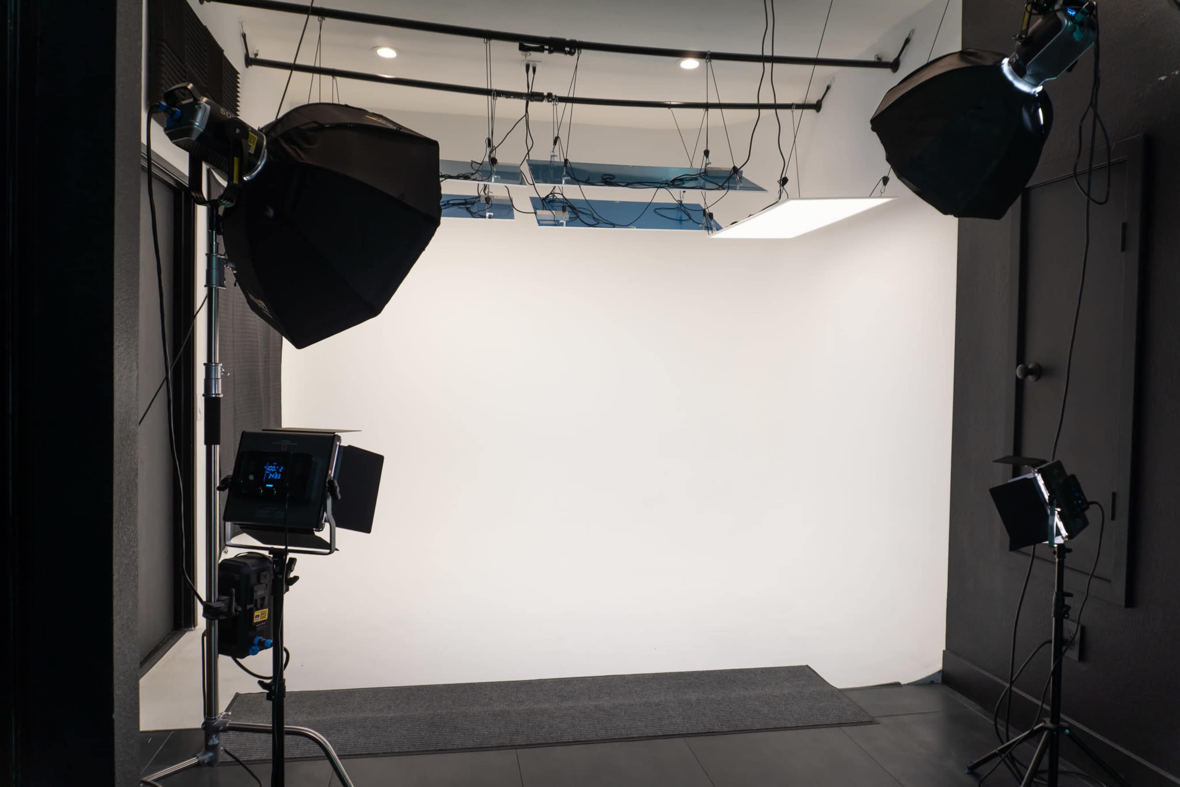 The image shows a photography studio with a white backdrop, equipped with several light sources and softboxes.