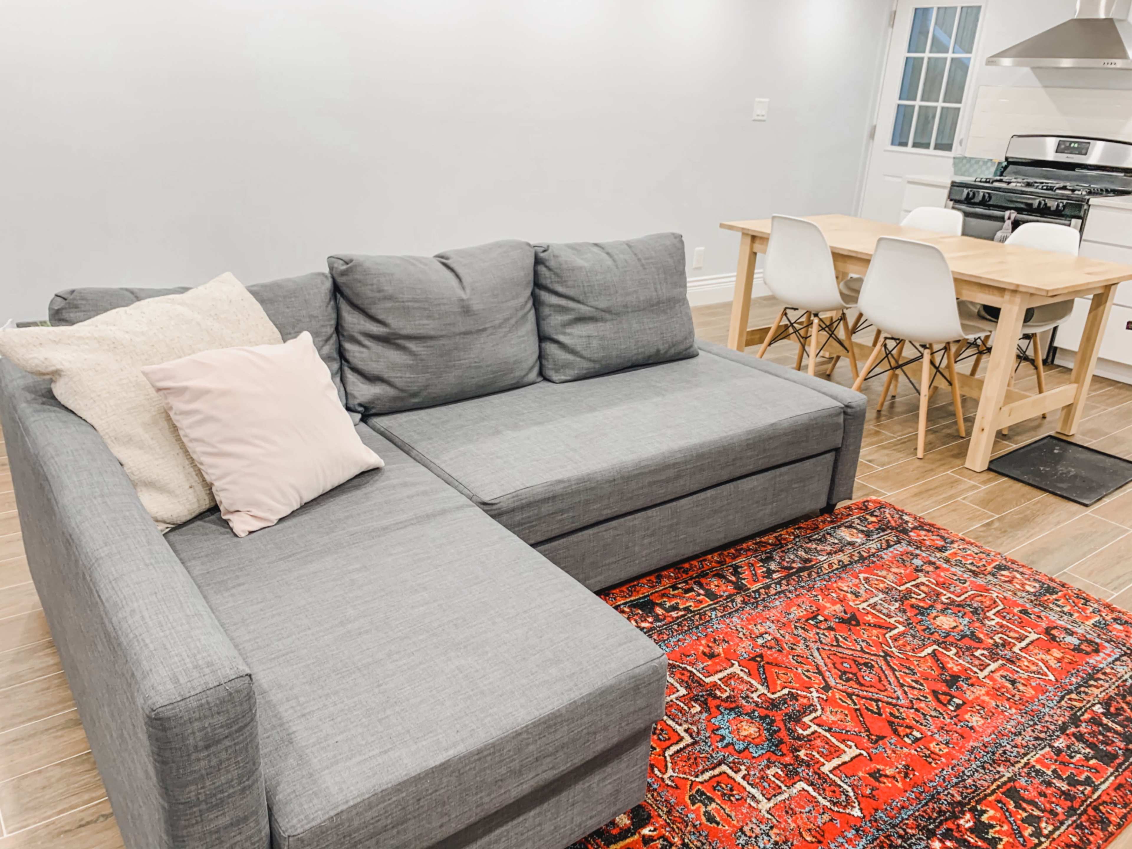 A gray sectional sofa with pillows, a red patterned rug, a wooden dining table with white chairs, and a kitchen area in the background.