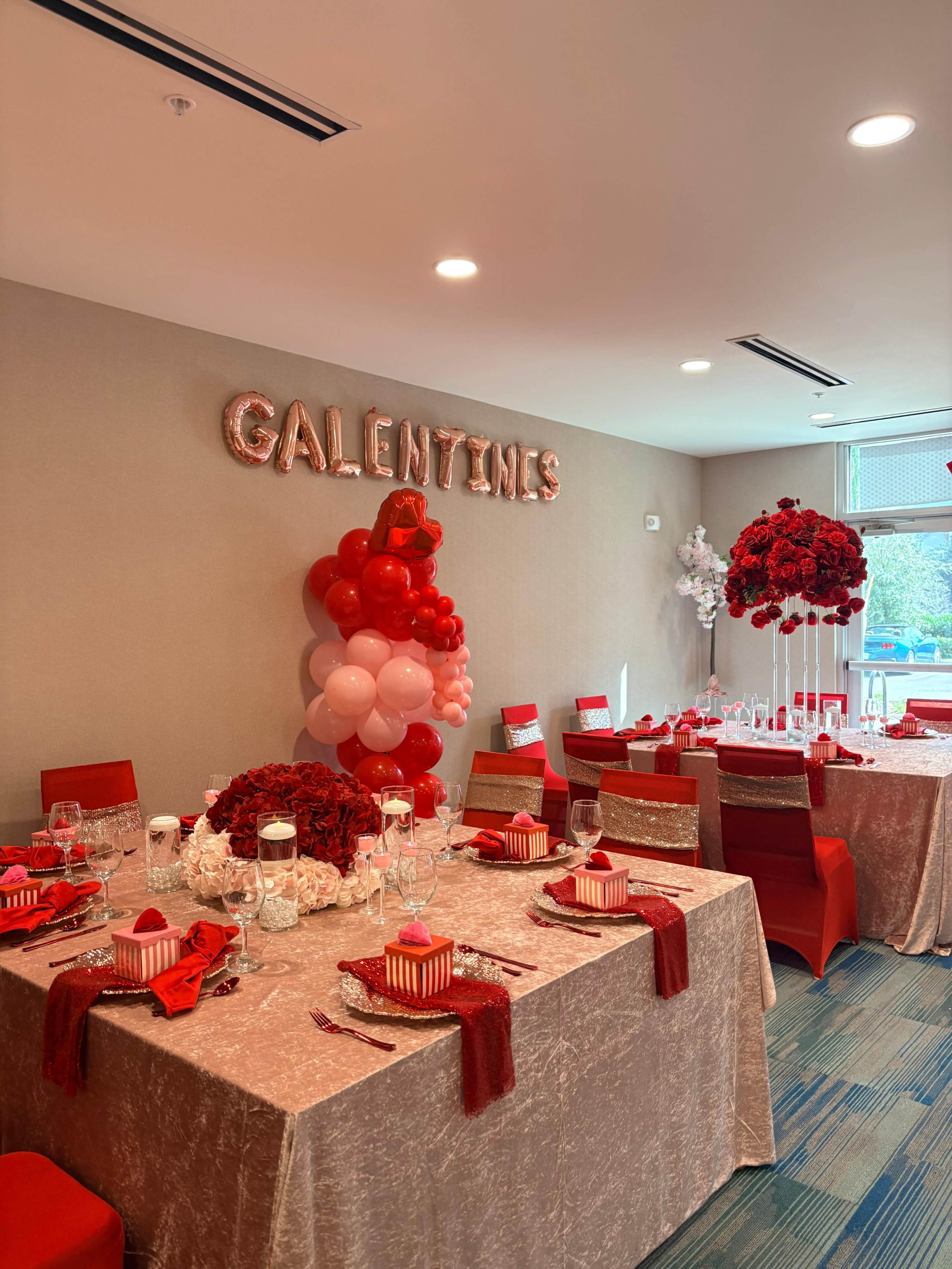 A decorated room features tables set for a Galentine's celebration, adorned with red and pink balloons, floral arrangements, and themed décor.