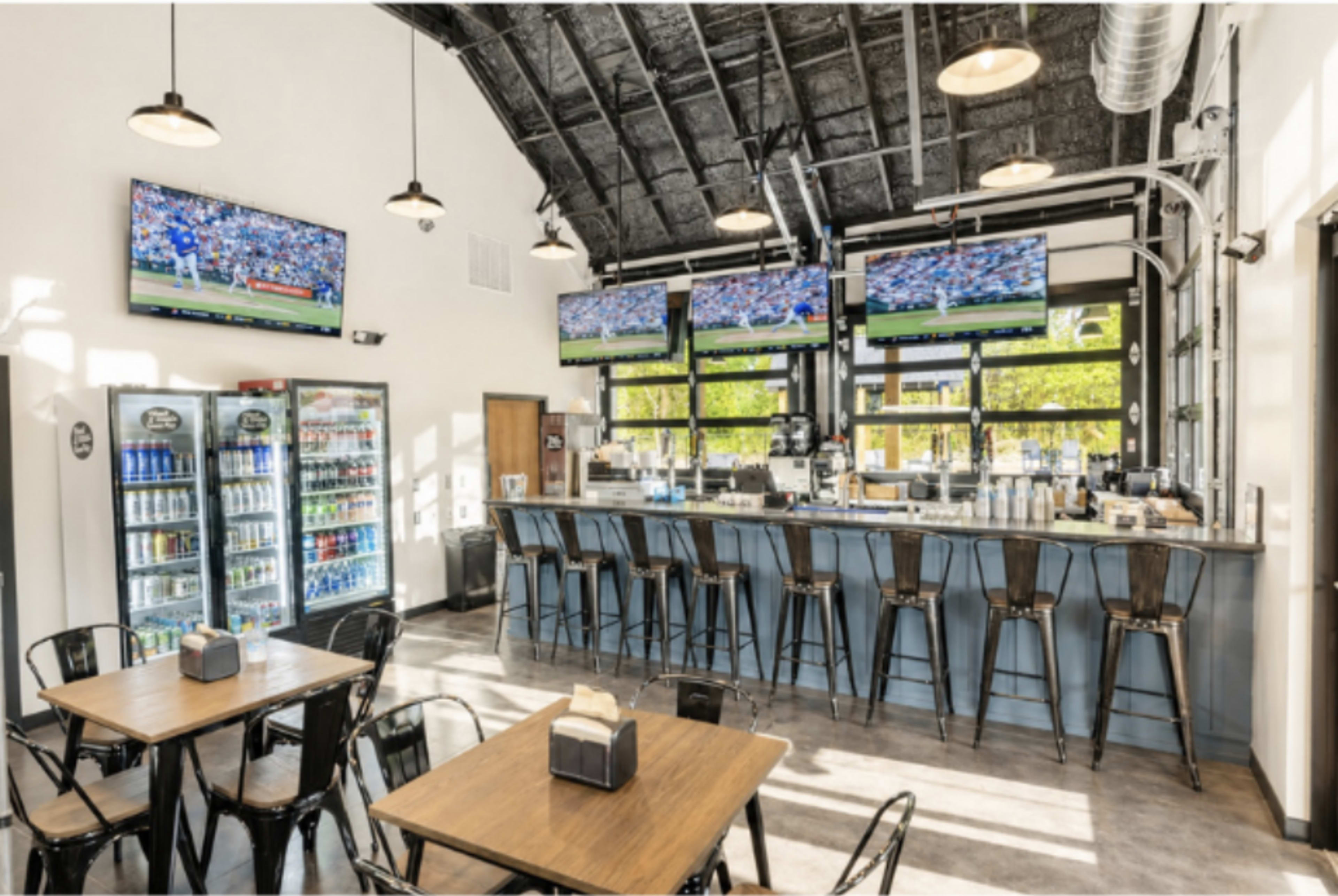 The image depicts a modern bar and dining area with four flat-screen TVs mounted on the wall, a beverage cooler, and several tables and chairs arranged on the floor.