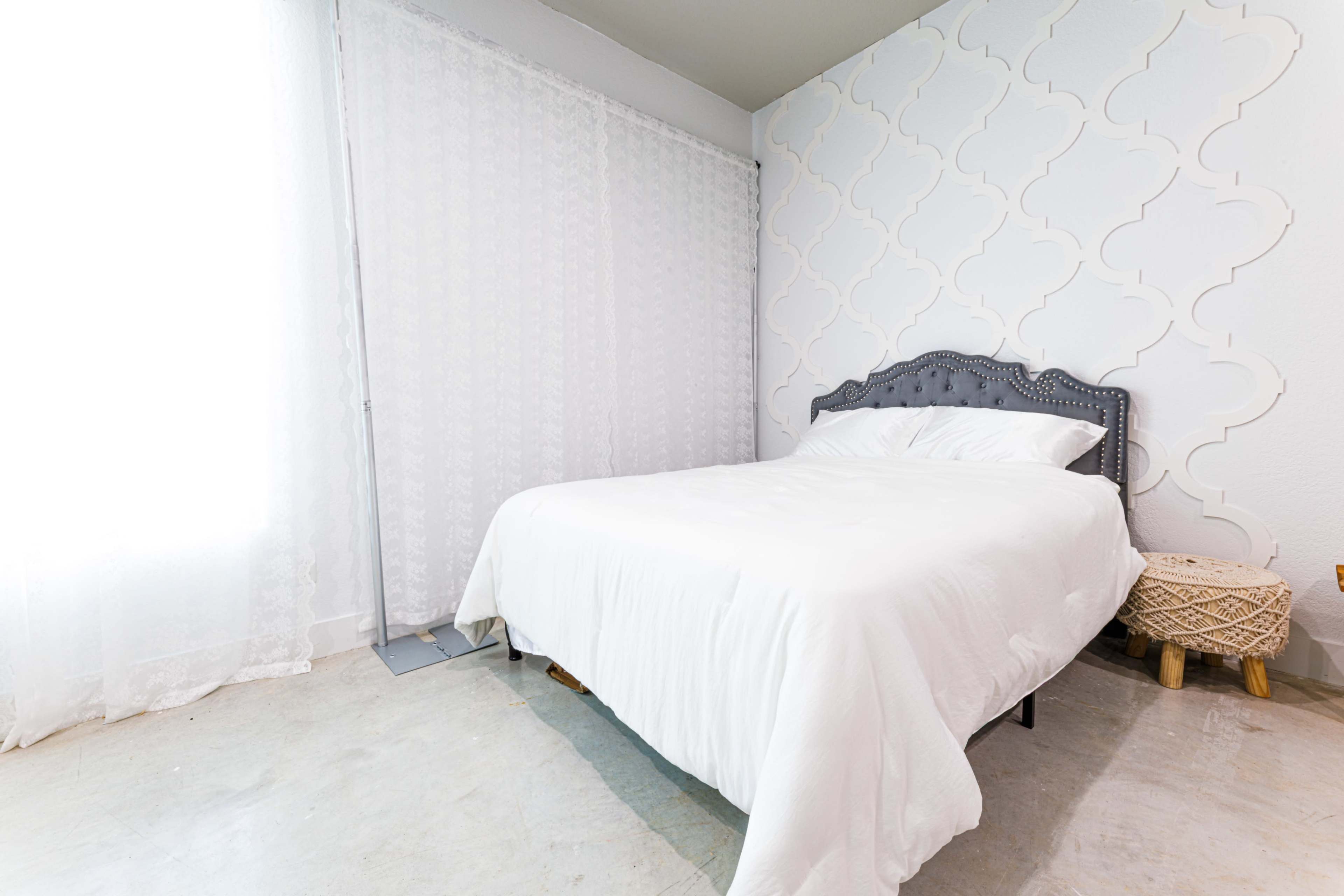 A simple bedroom features a white bed with a patterned headboard, light curtains, and a decorative stool beside it.