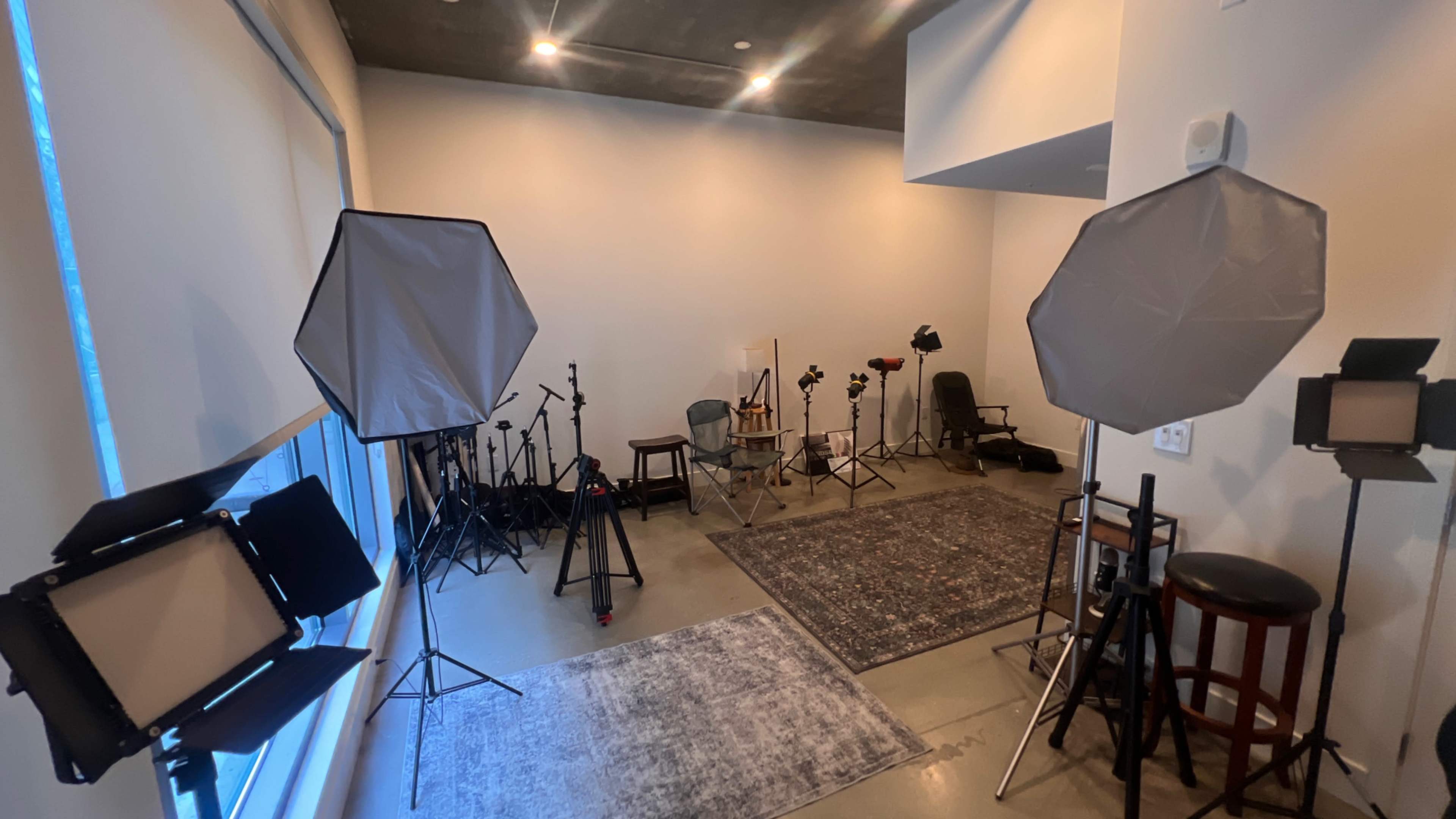 The image shows a studio space equipped with various photography and lighting equipment, including softboxes and tripods, arranged around a central area with a rug and seating.