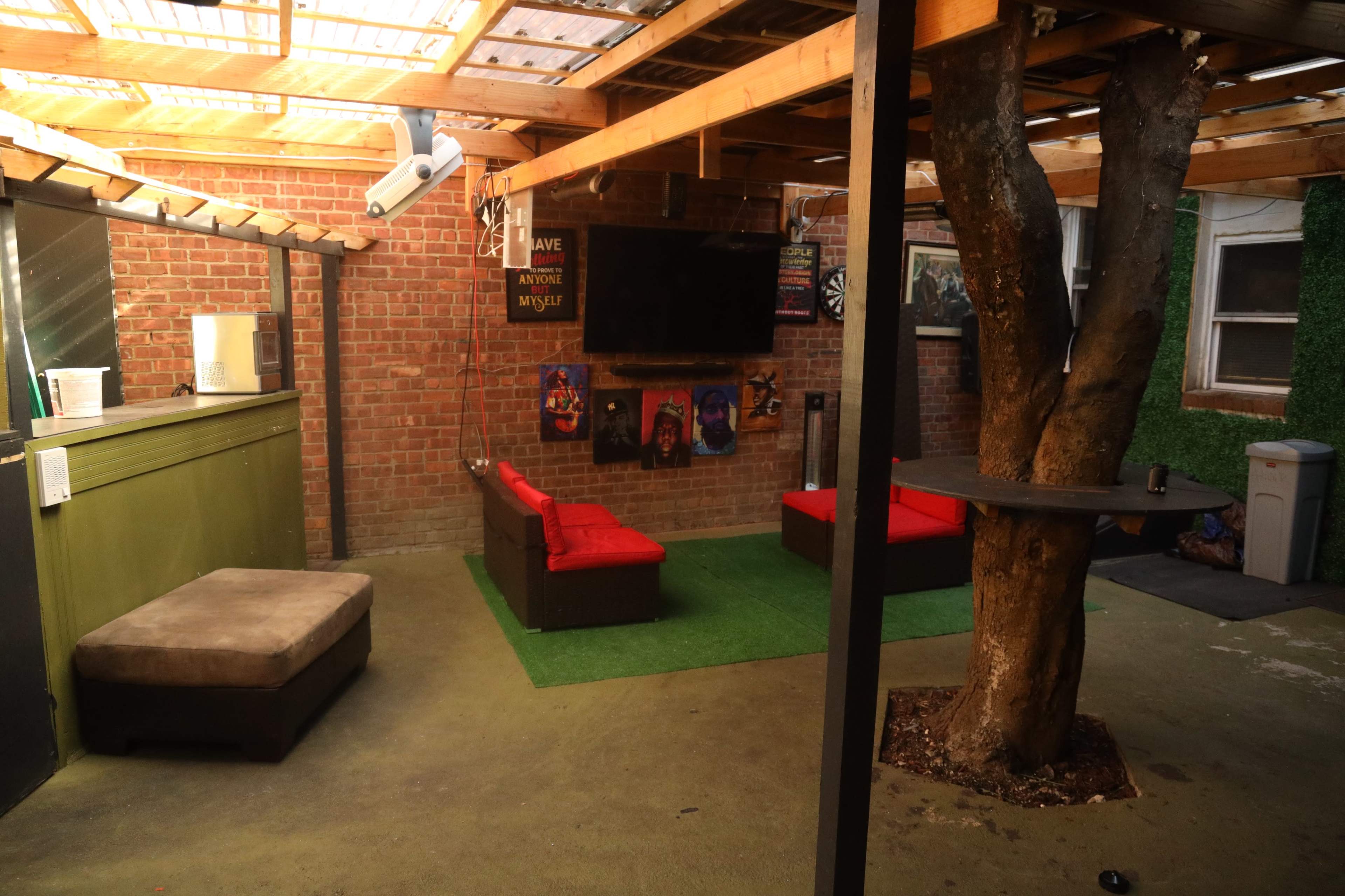A spacious indoor area with a brick wall, a flat-screen TV mounted on the wall, red seating, a tree in the center, and green carpeting on the floor.