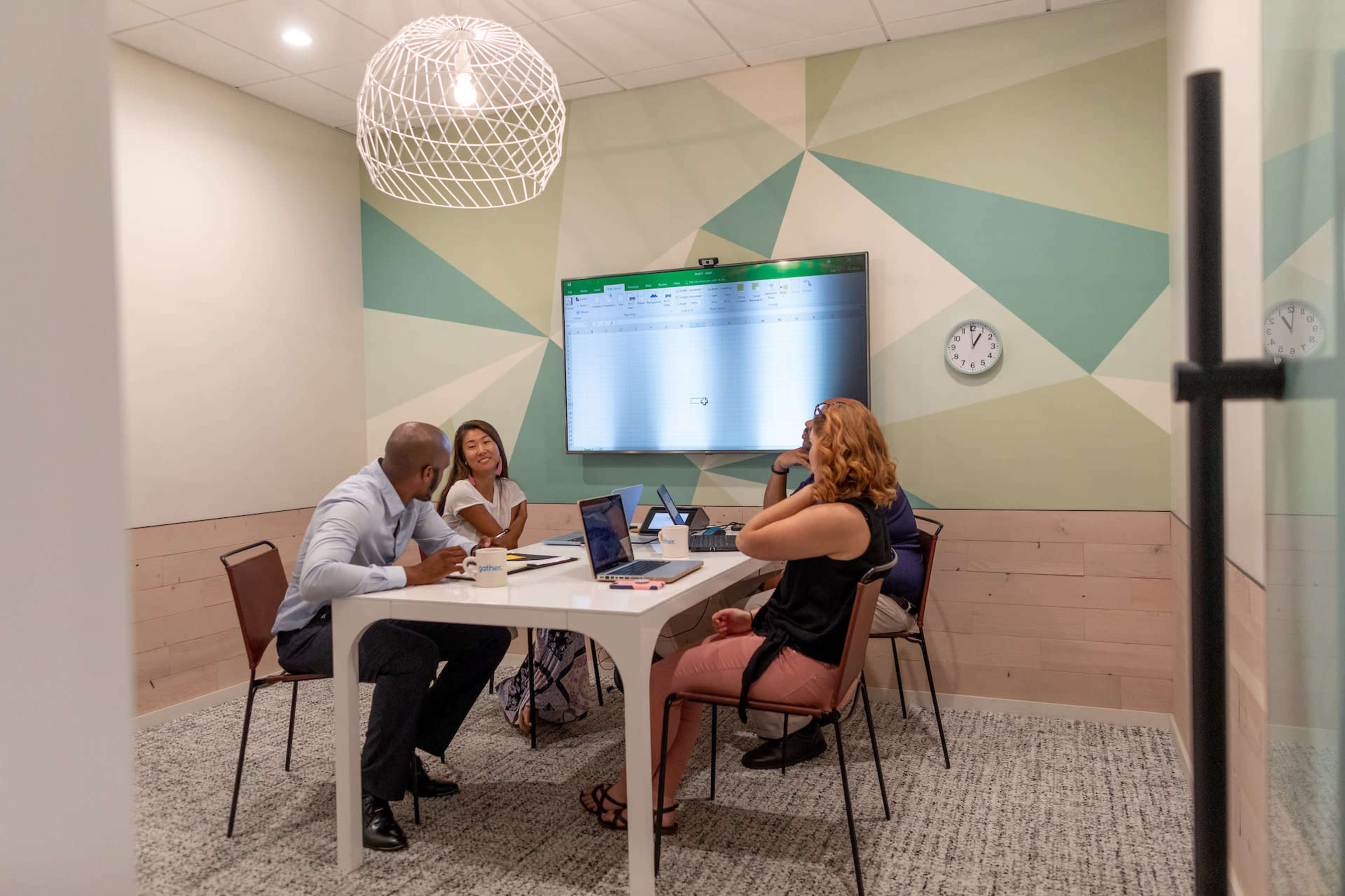 A group of four people is engaged in a discussion around a table in a small meeting room with a large screen displaying a spreadsheet.