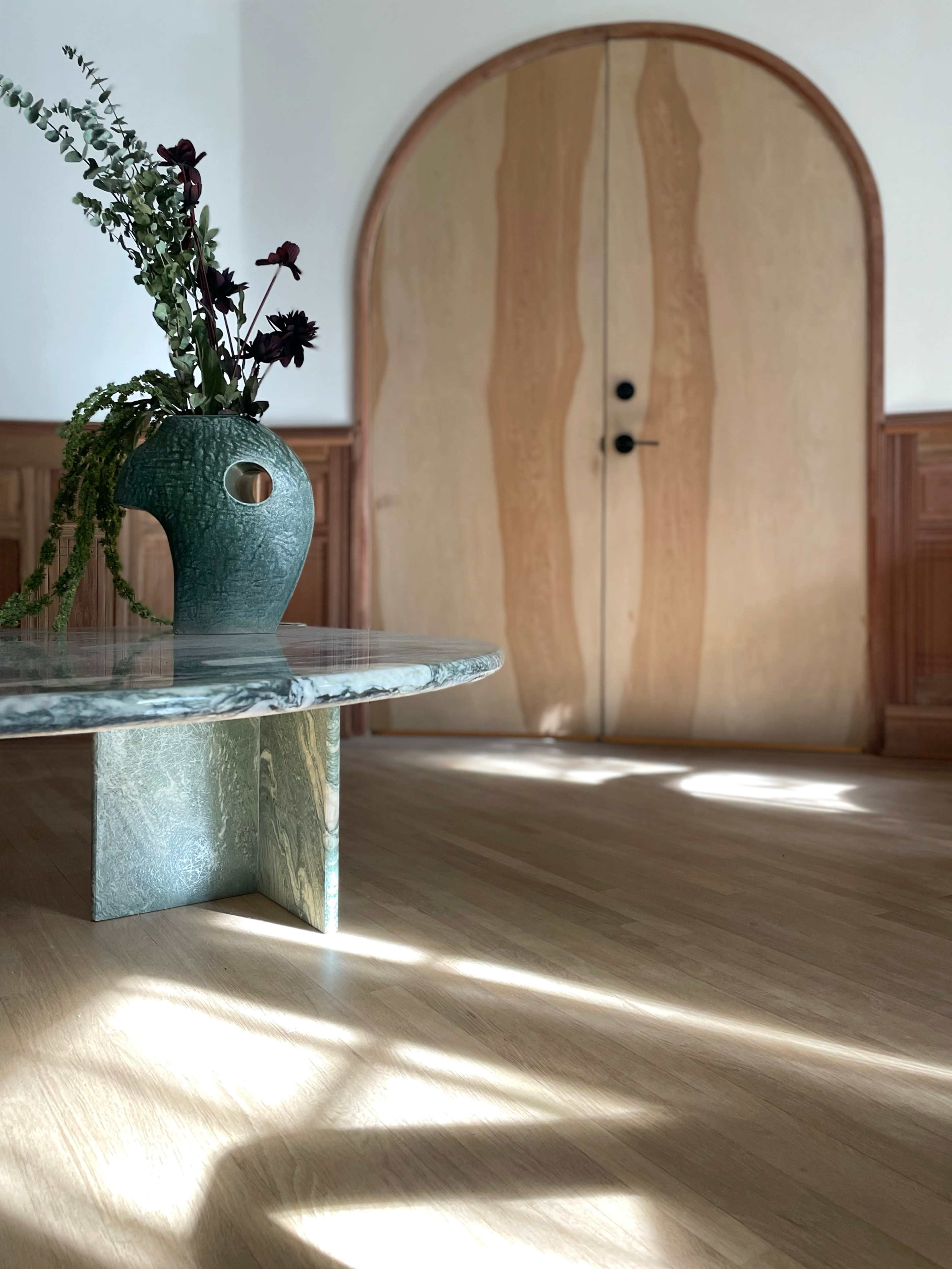 A modern room features a circular table with a glass top and a textured green vase holding flowers, complemented by an arched wooden door in the background.