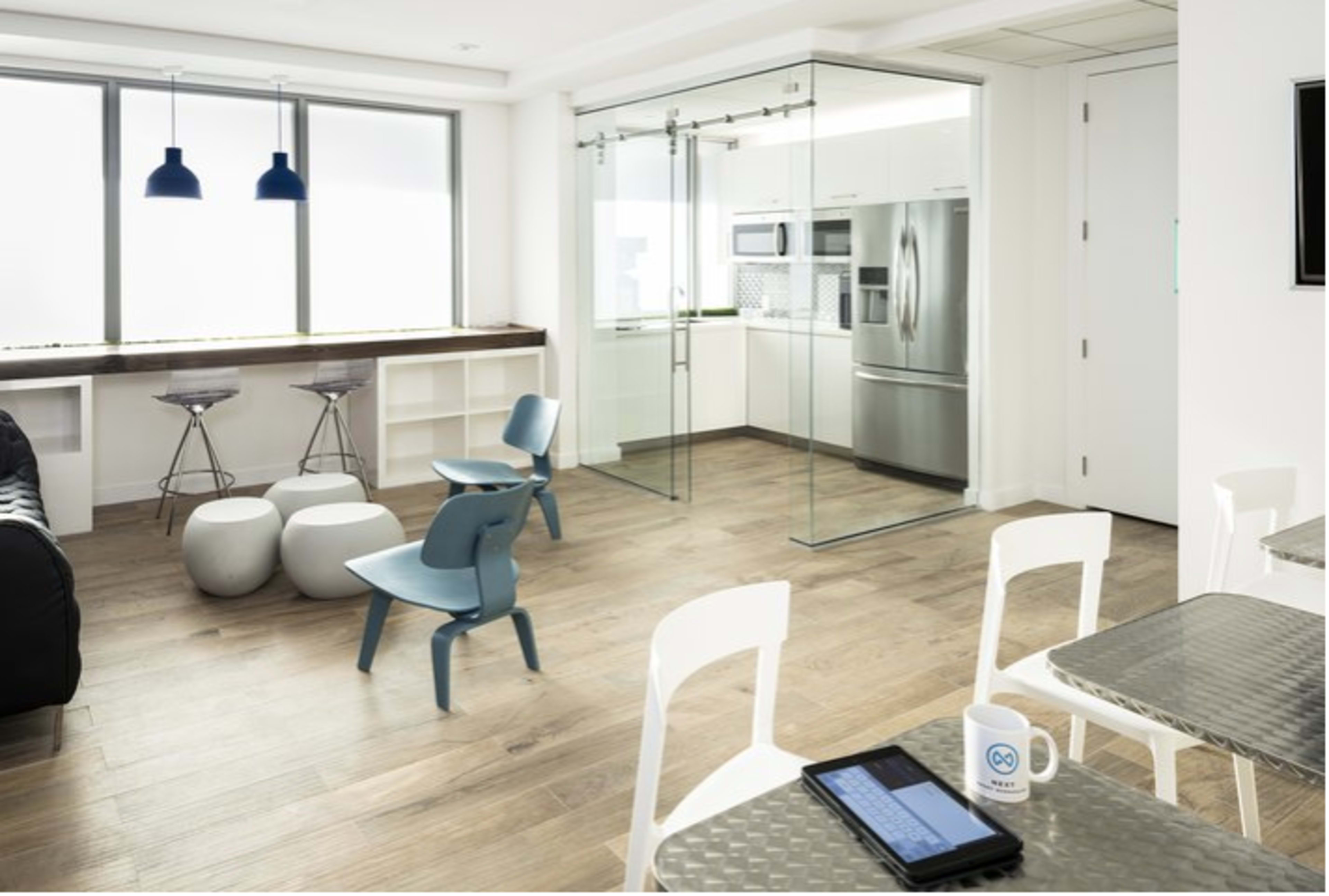 A modern interior features a kitchen with stainless steel appliances, a glass-walled area, and seating arrangements with stools and chairs on a wooden floor.