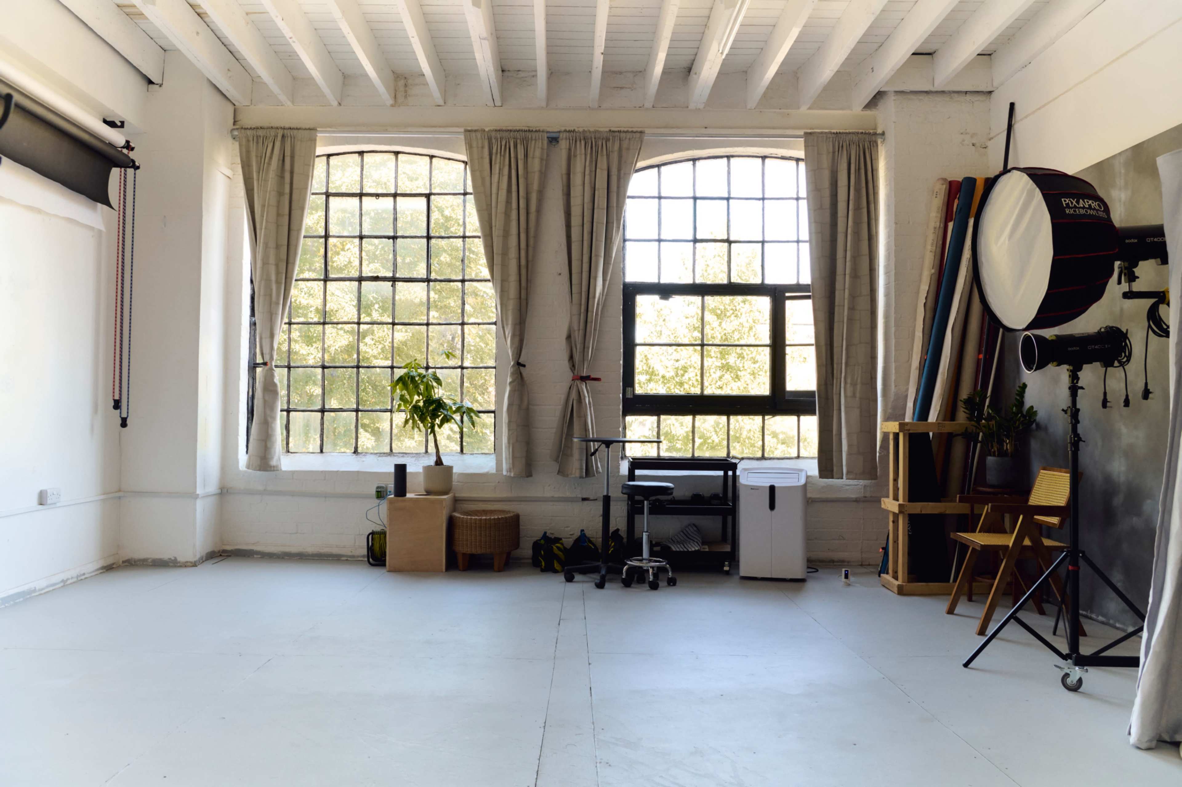 The image shows a spacious photo studio with large windows, natural light streaming in, and various photography equipment along one wall.
