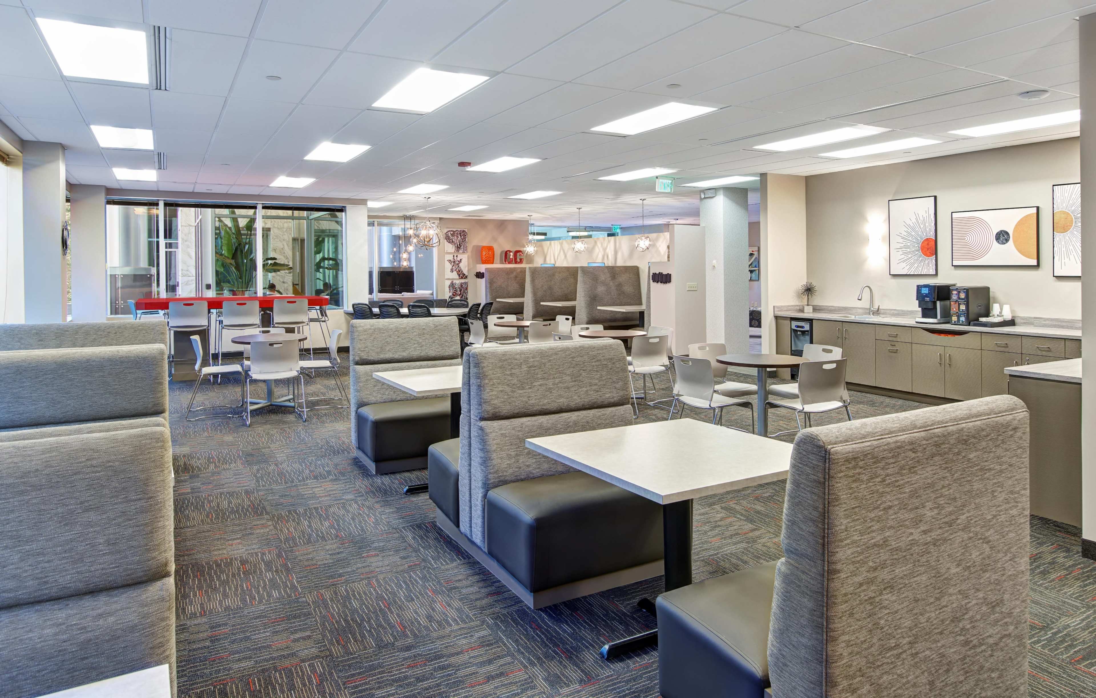 The image shows a modern office break area with seating booths, tables, and a kitchenette.
