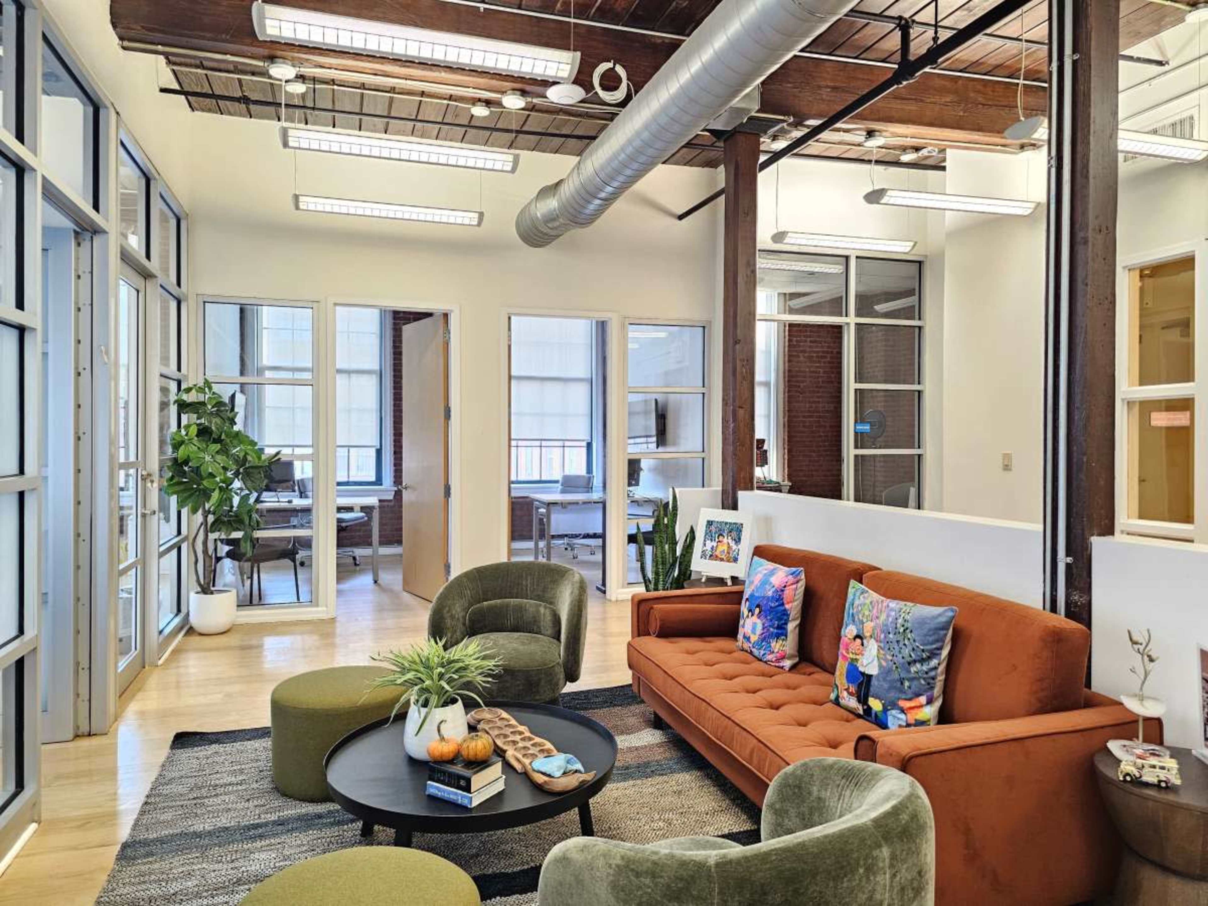 The image shows a modern office space with a vibrant orange sofa, green armchairs, a round coffee table, and large windows allowing natural light to enter.