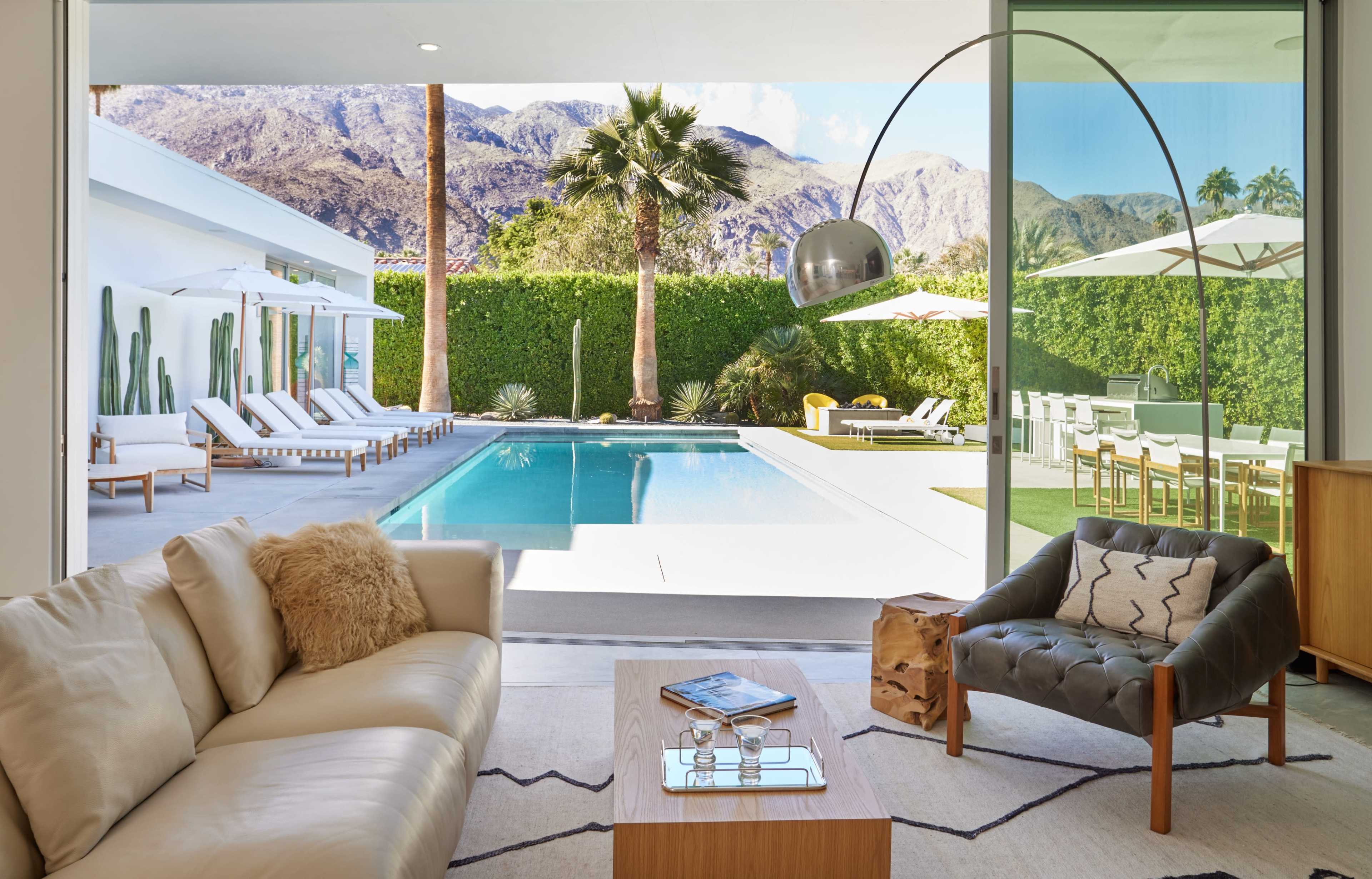 The image shows a modern living room with a light-colored sofa, a coffee table, and a view of a pool surrounded by palm trees and mountains through large glass doors.