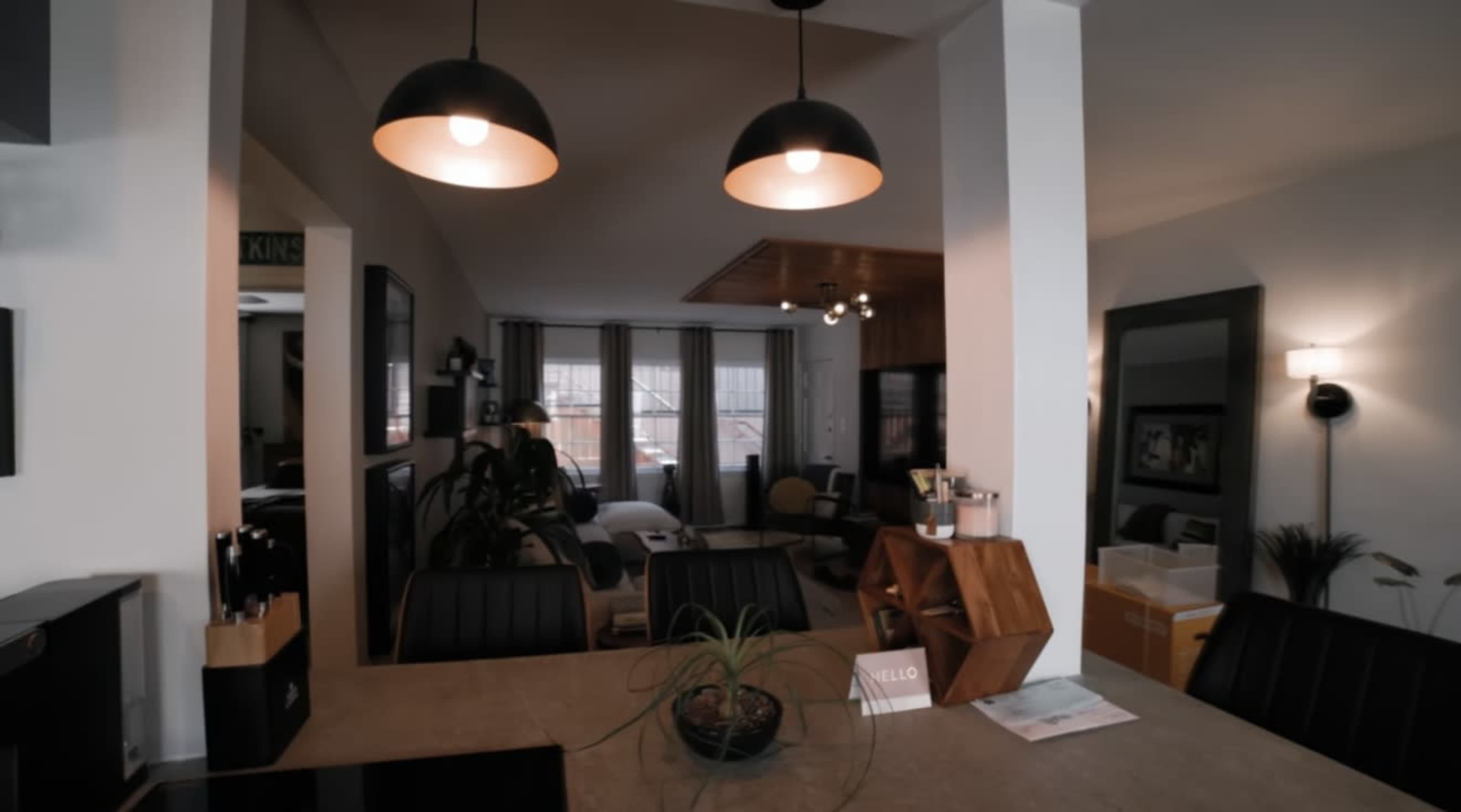 The image shows a modern interior space with a dining area featuring a stone countertop and two pendant lights, leading to a living room area with large windows.