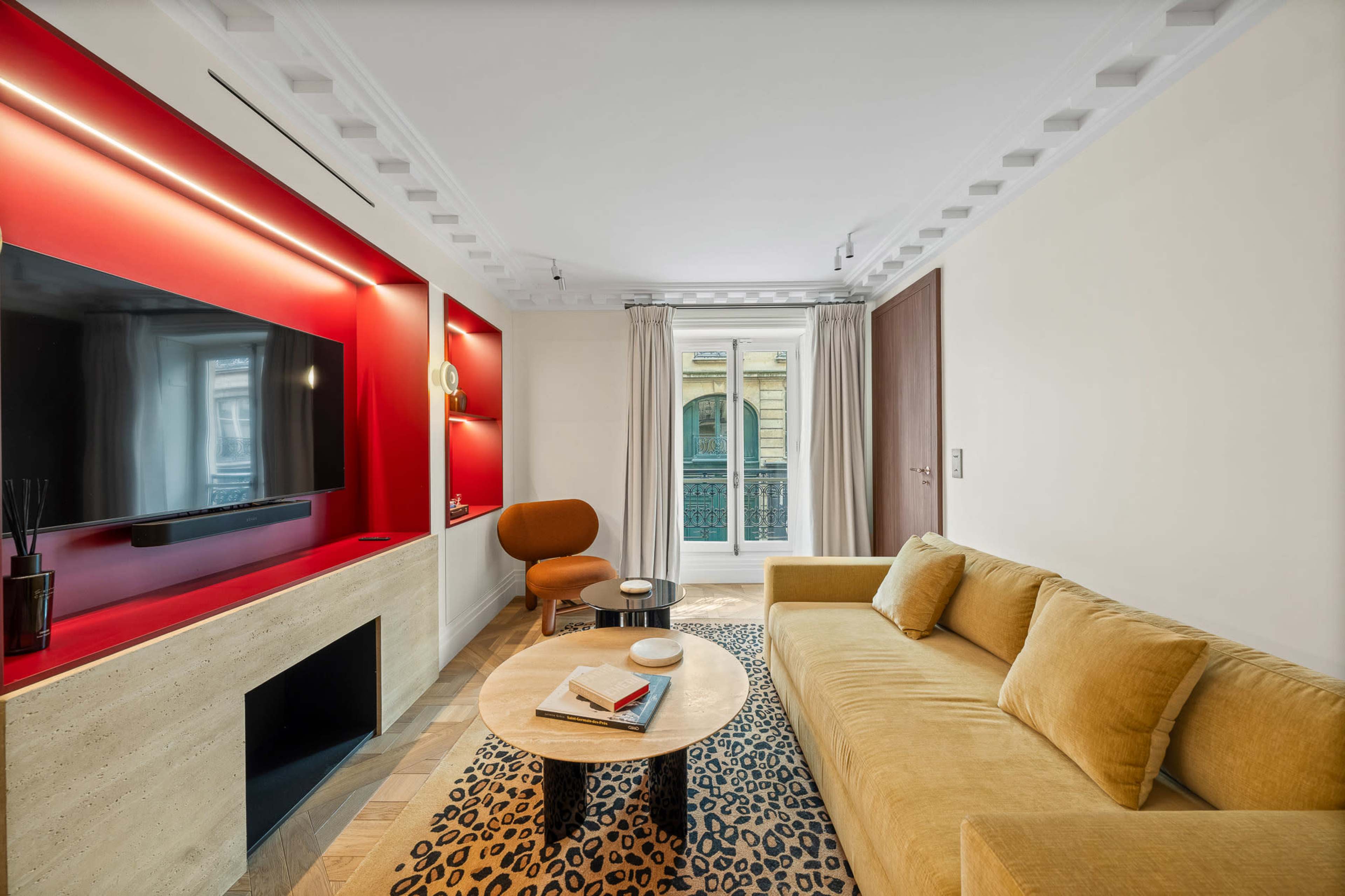 The image shows a modern living room with a beige sofa, a round wooden coffee table on a patterned rug, a large television mounted on a red accent wall, and a balcony visible through the window.